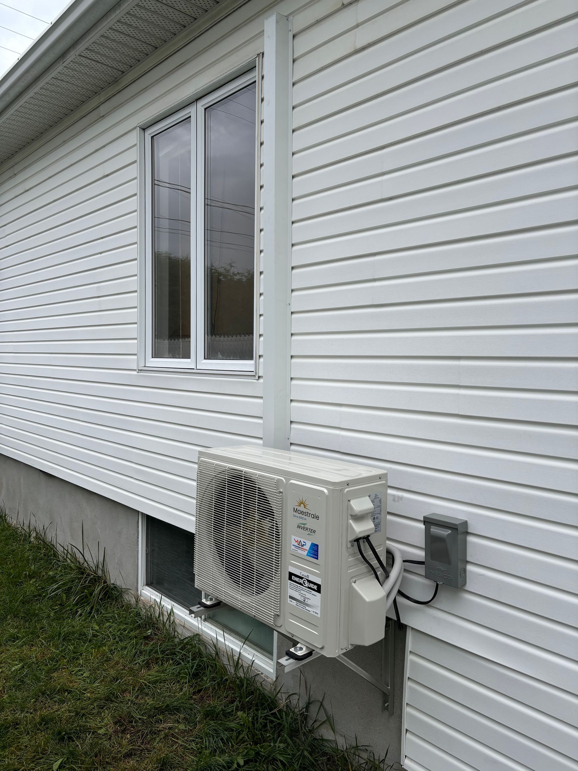 Unité de climatisation blanche fixée sur la façade d'une maison blanche avec fenêtre et pelouse.