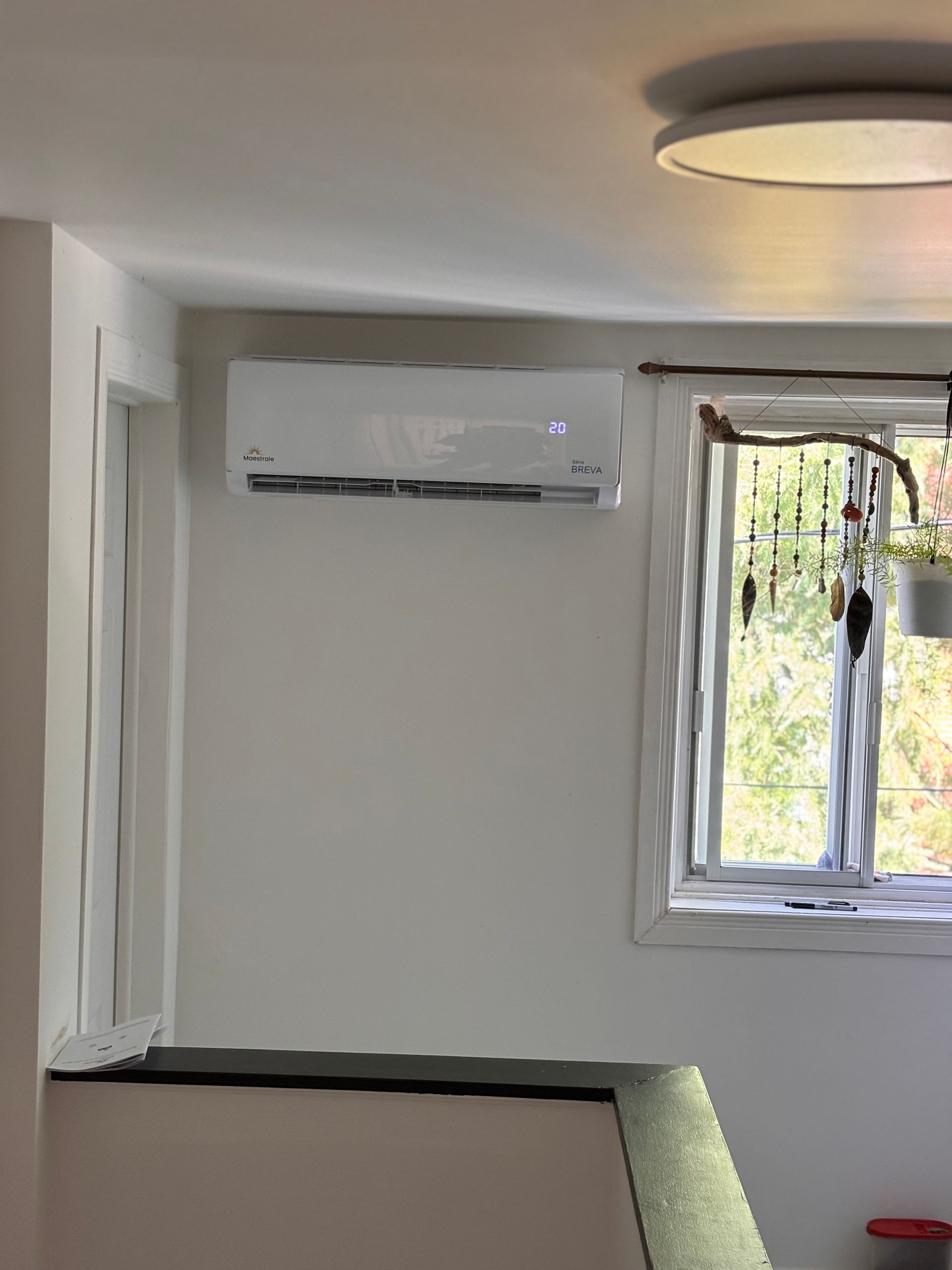 White wall-mounted air conditioner installed in a bedroom with window and hanging plant.