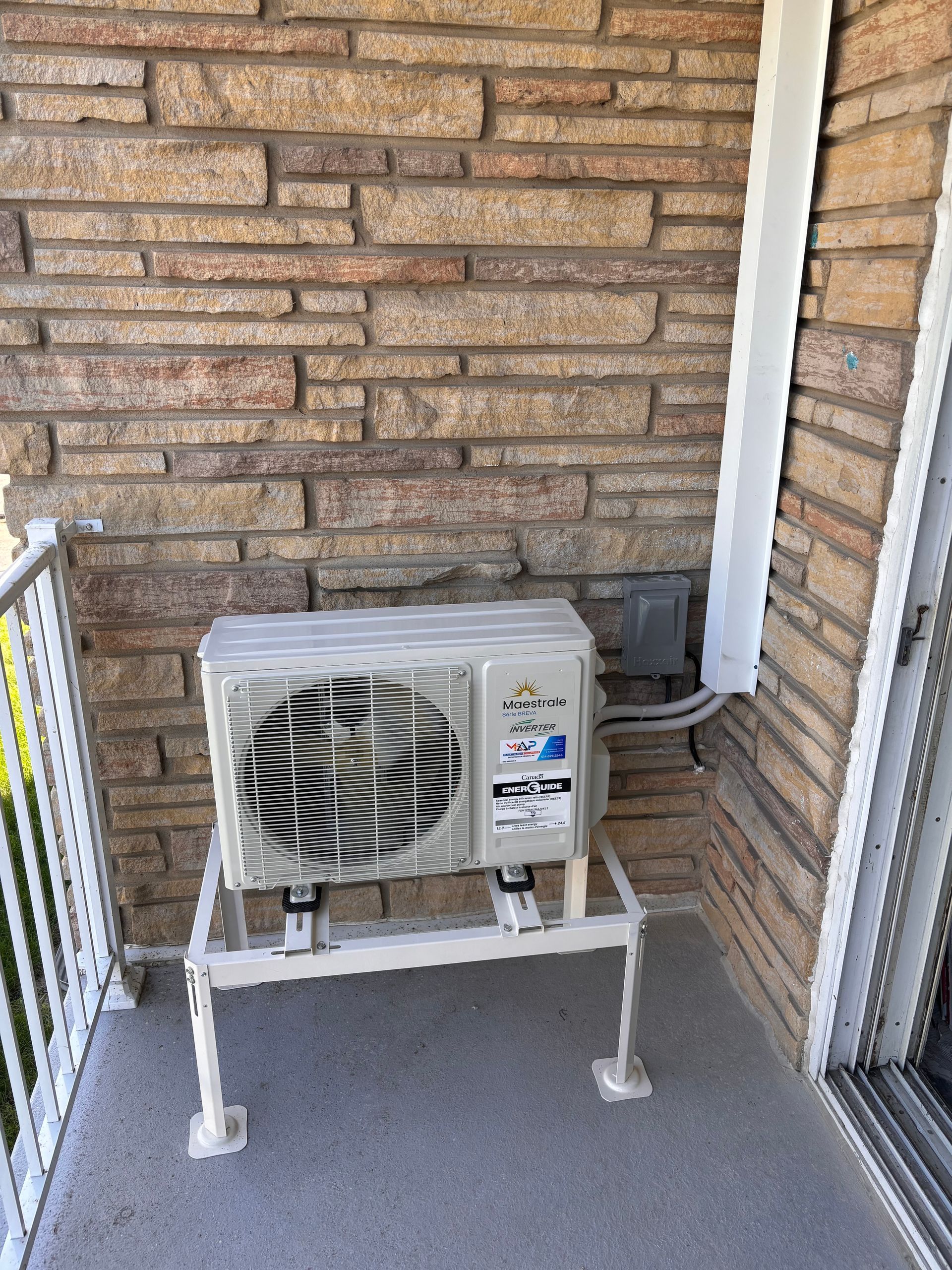 Unité de climatisation sur un support en métal blanc, fixée sur un balcon en pierre à côté d'une rambarde blanche.