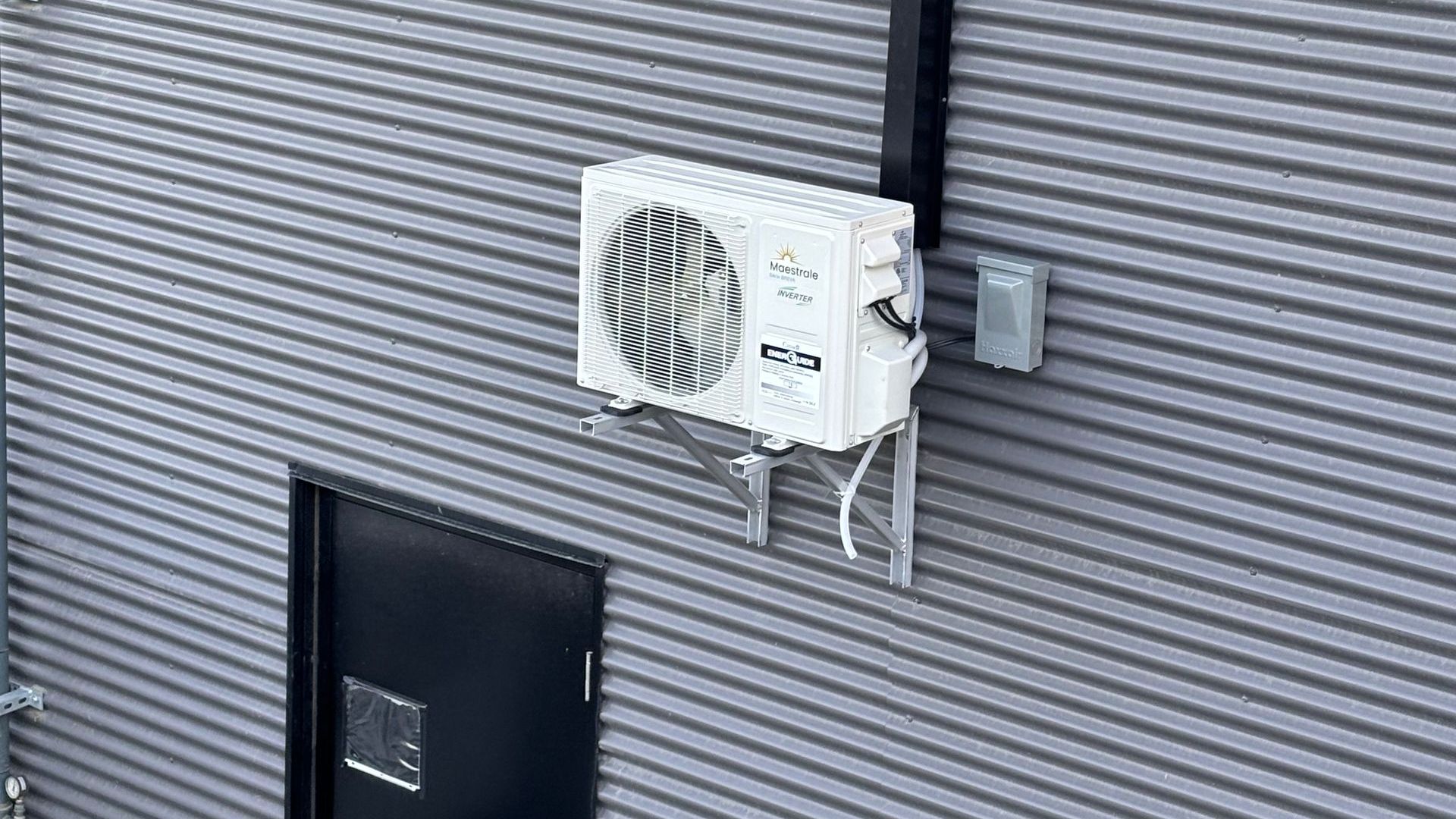 Unité de climatisation blanche fixée sur un mur en tôle ondulée grise, près d'une porte sombre.
