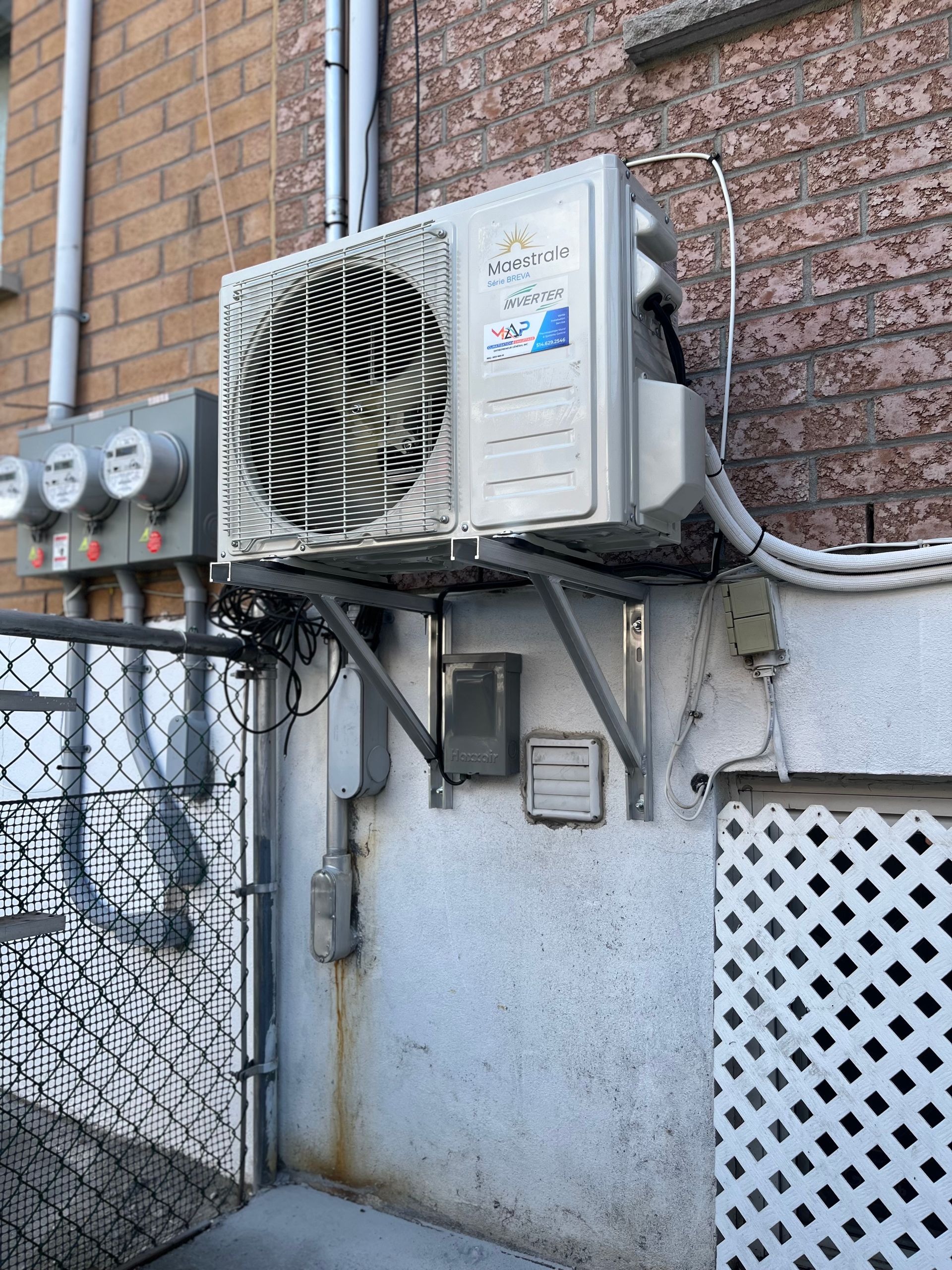 Unité de climatisation extérieure fixée sur le mur d'un bâtiment, près d'une clôture en grillage et de compteurs électriques.