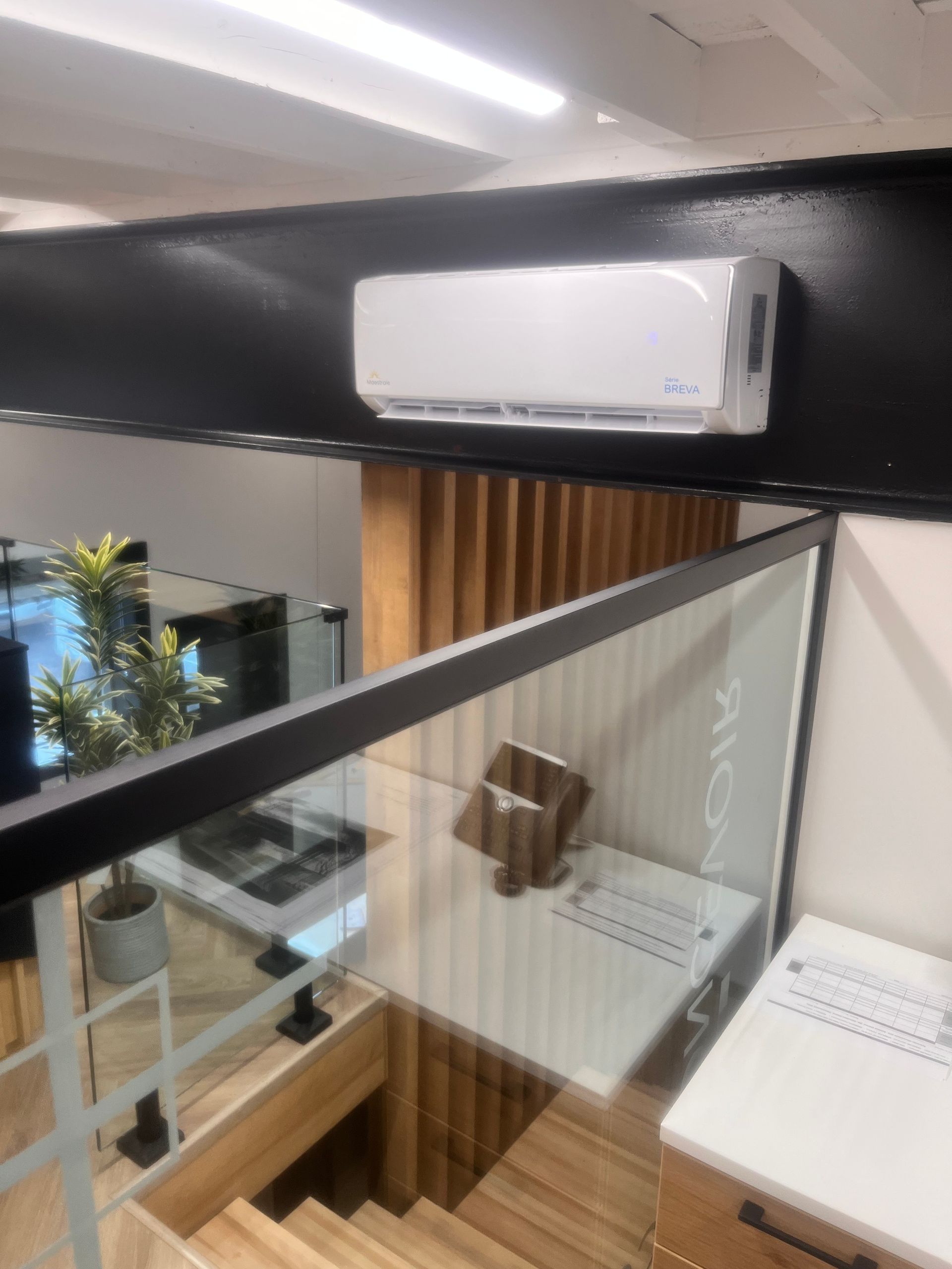 Unité de climatisation fixée sur un mur noir au-dessus d'un escalier avec une rampe en verre et des marches en bois.