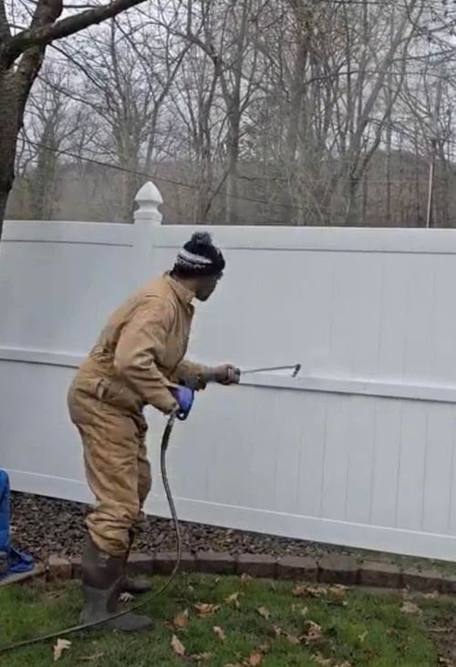 Person pressure-washing a white fence in a wooded yard, wearing tan work clothes and boots.