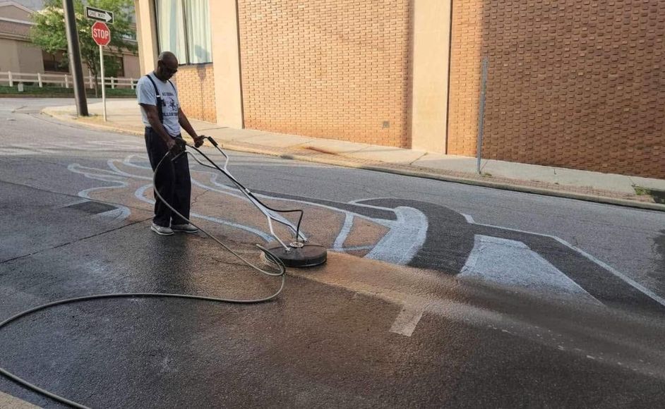 Worker cleaning street with pressure washer on wet pavement beside a brick wall.