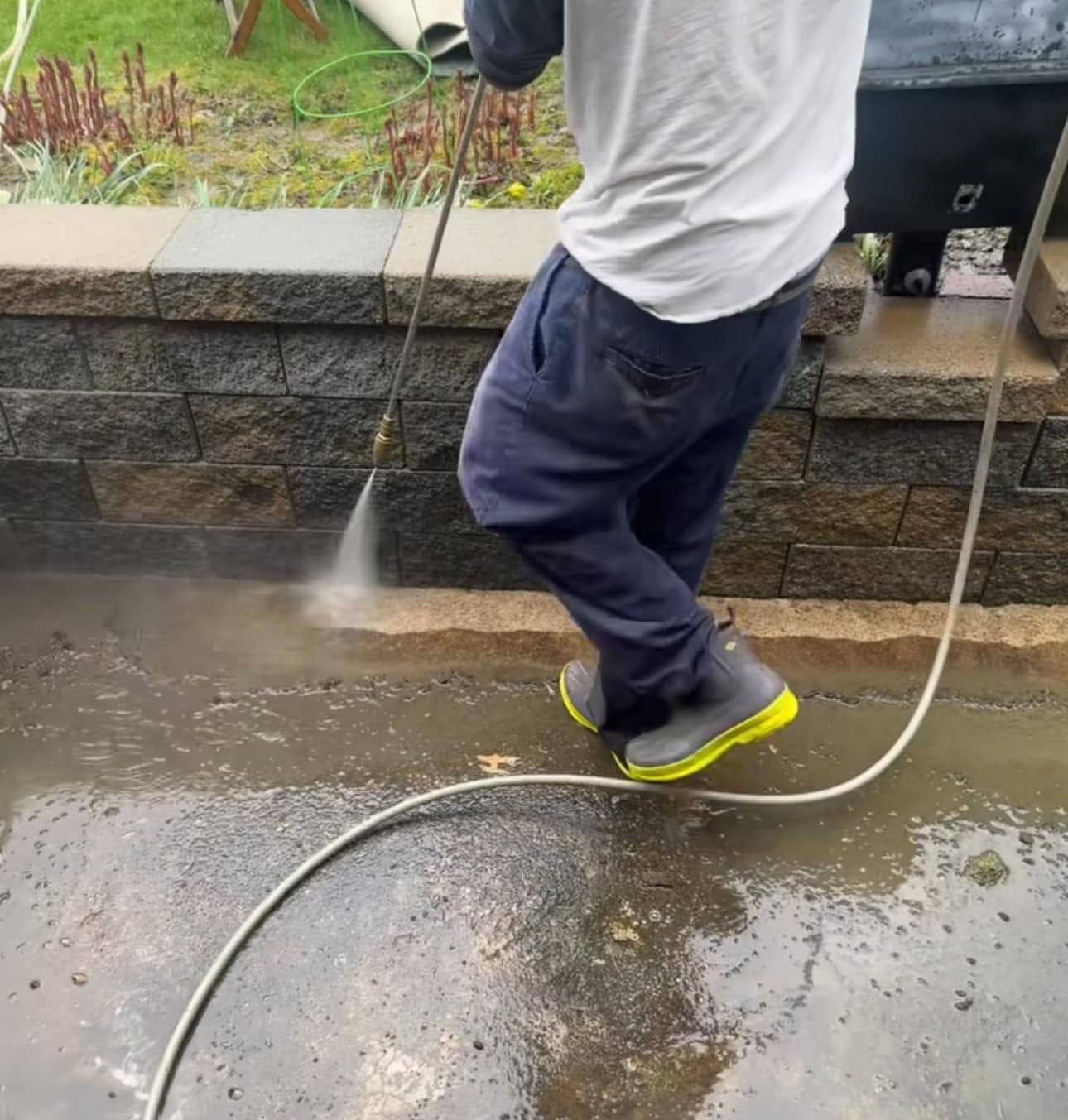 Person pressure washing a wet concrete patio beside a stone retaining wall.