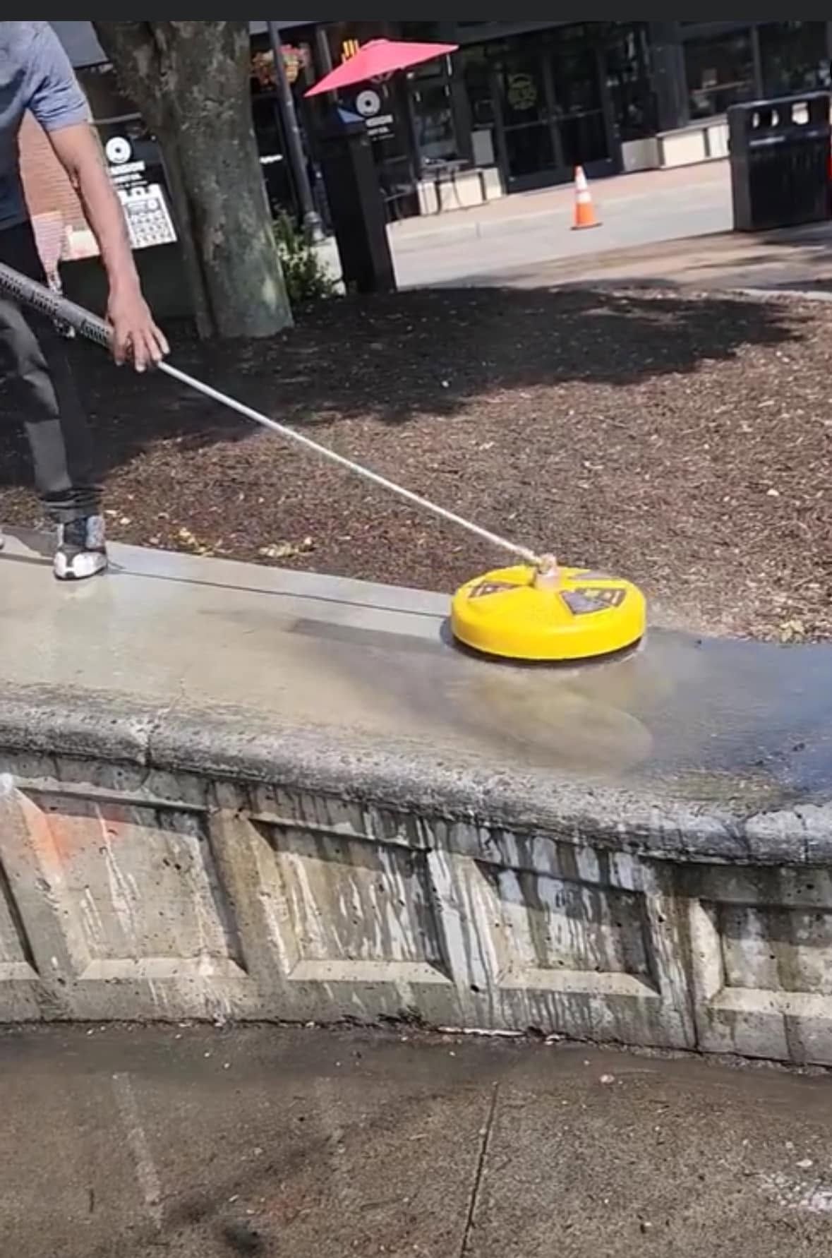 Person using a yellow edge-cleaning tool on a curb in an outdoor plaza area