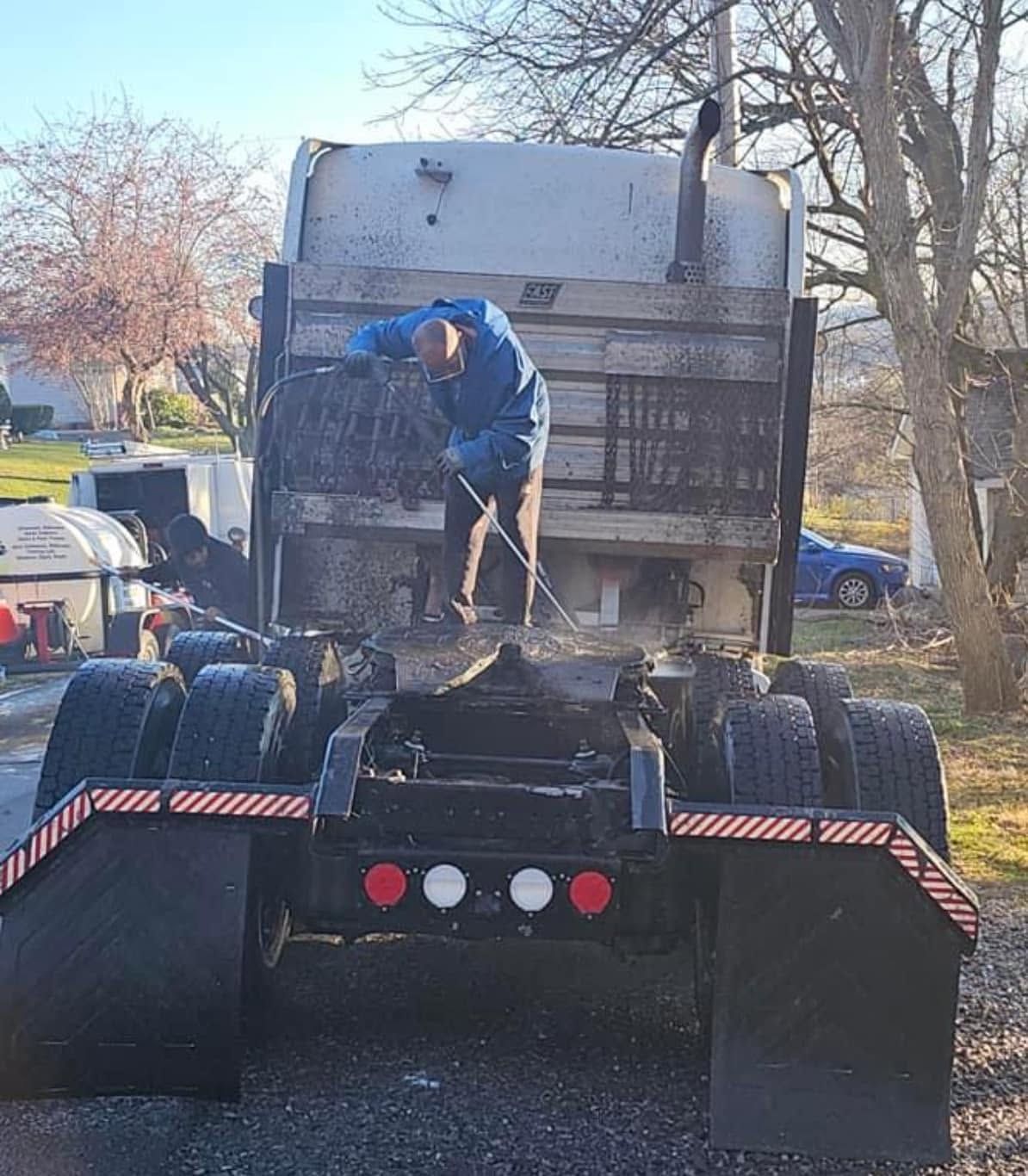 Worker on a truck bed using a pressure washer to clean a large truck outdoors.