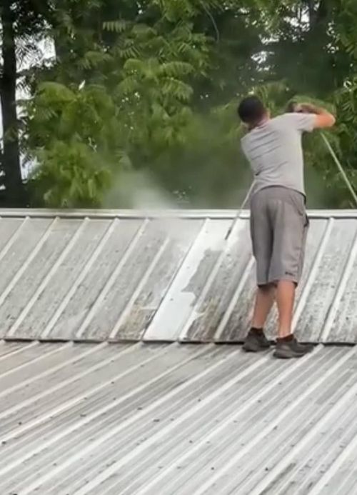 Man on a metal roof spraying water toward the tree line near a house.