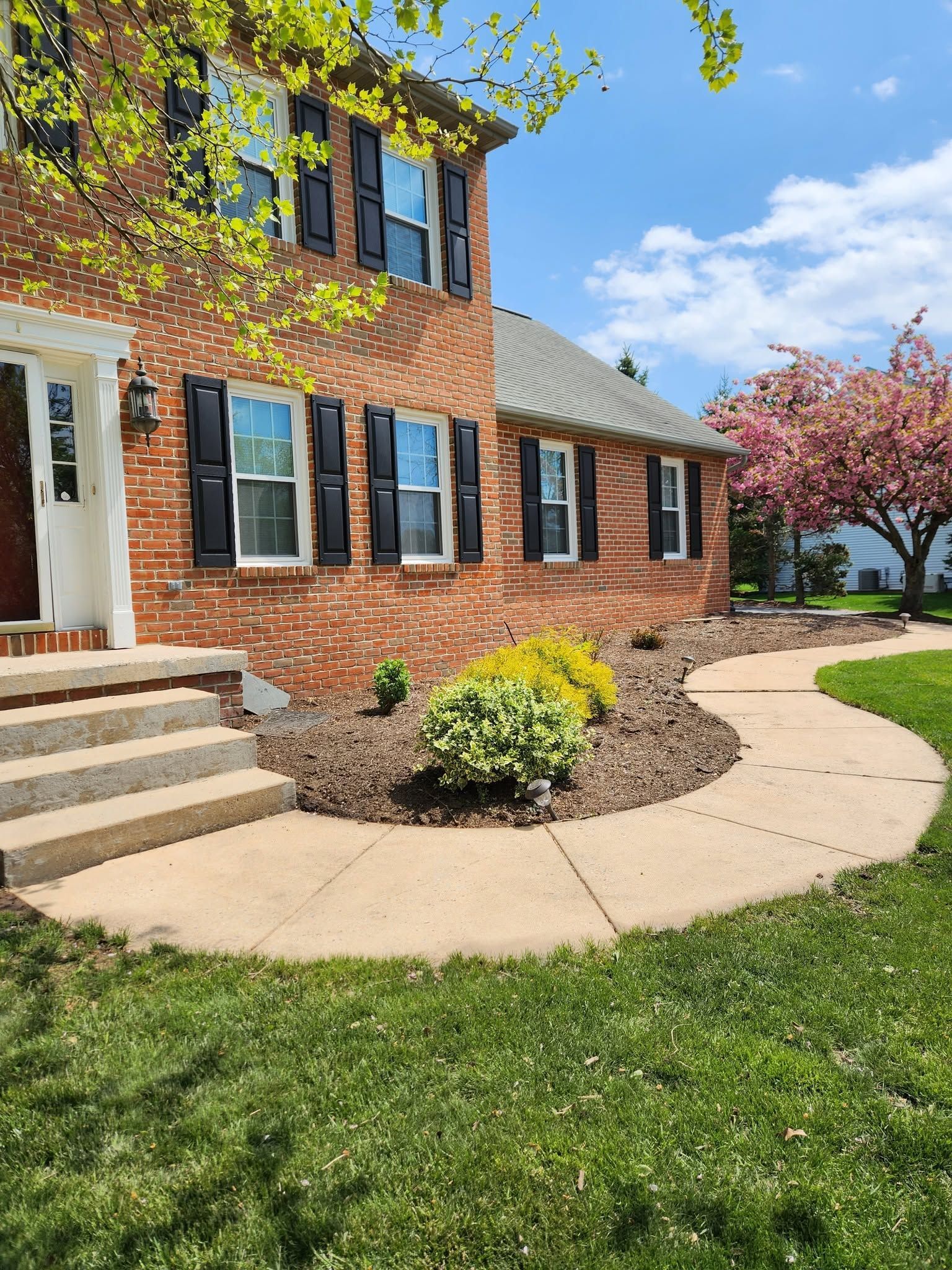 Brick house with black shutters, curved walkway, and green lawn under a blue sky