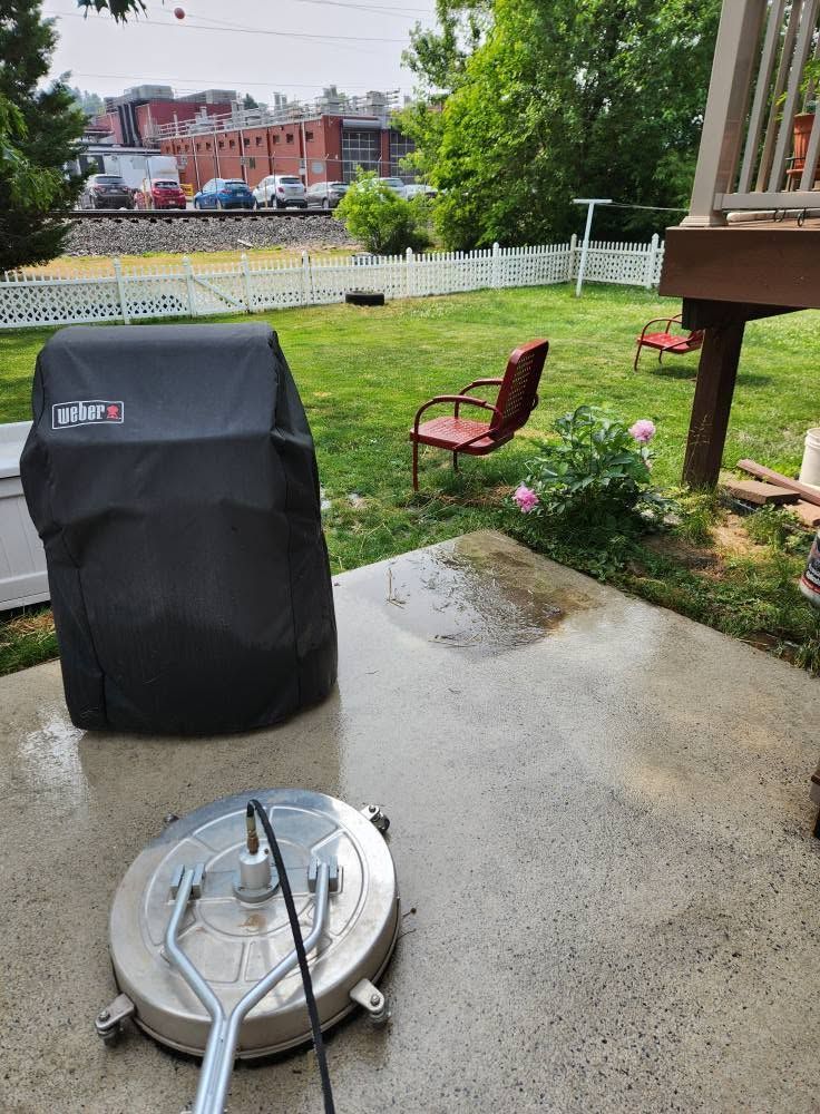 Rainy backyard patio with a covered grill, patio heater base, and two chairs near a fenced lawn.
