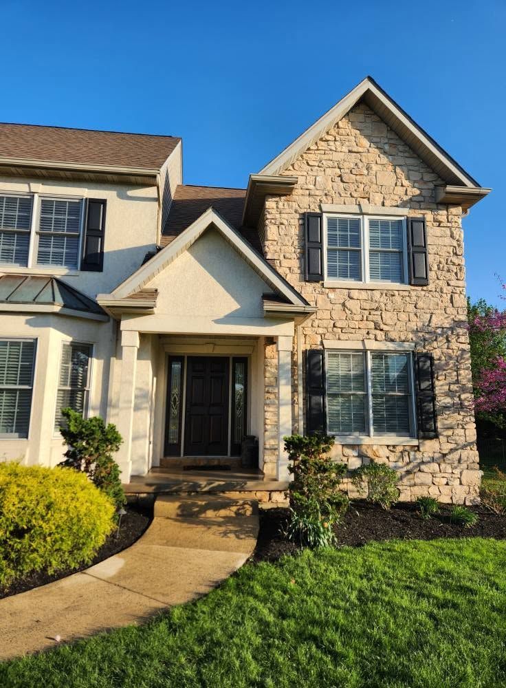 Two-story stone house with a front porch, walkway, and green lawn in bright daylight