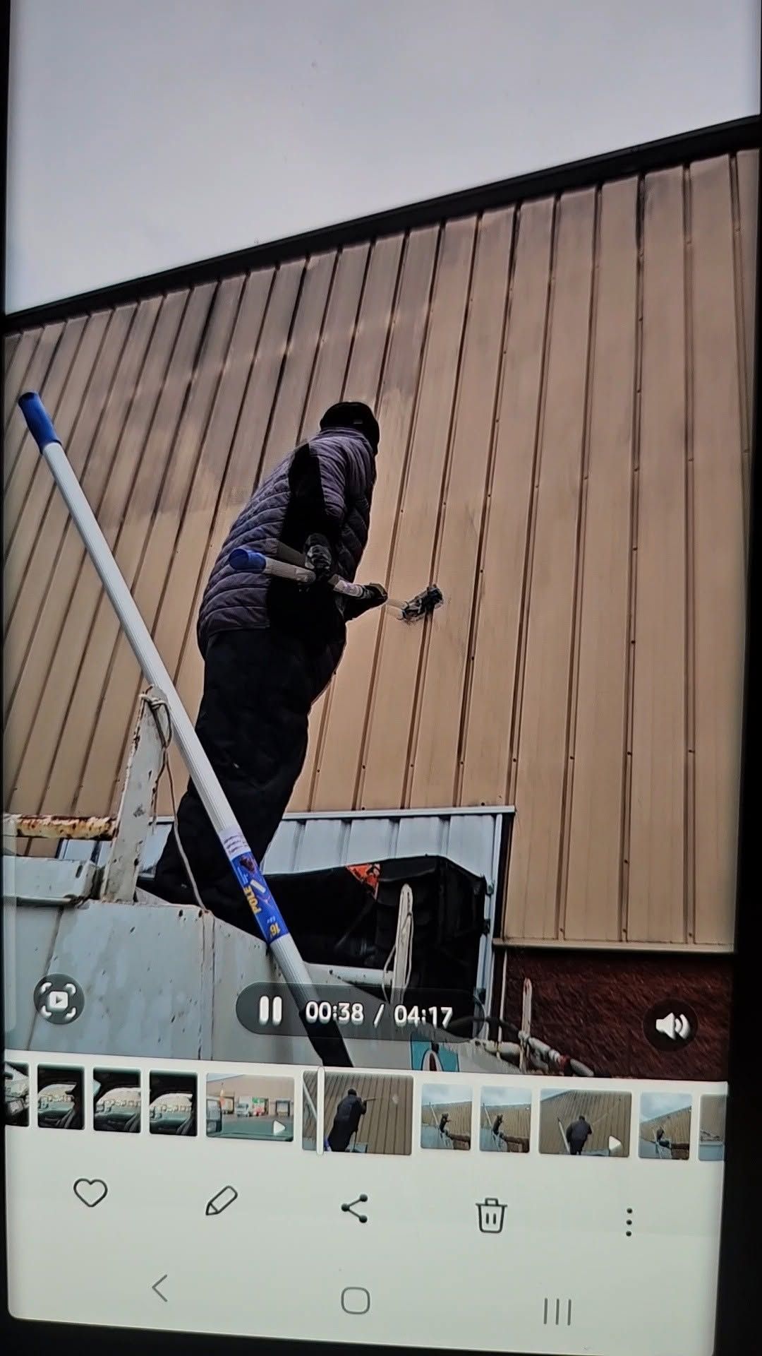 Person on a roof using a tool beside a white ladder, with a tan wooden wall and pale sky behind.