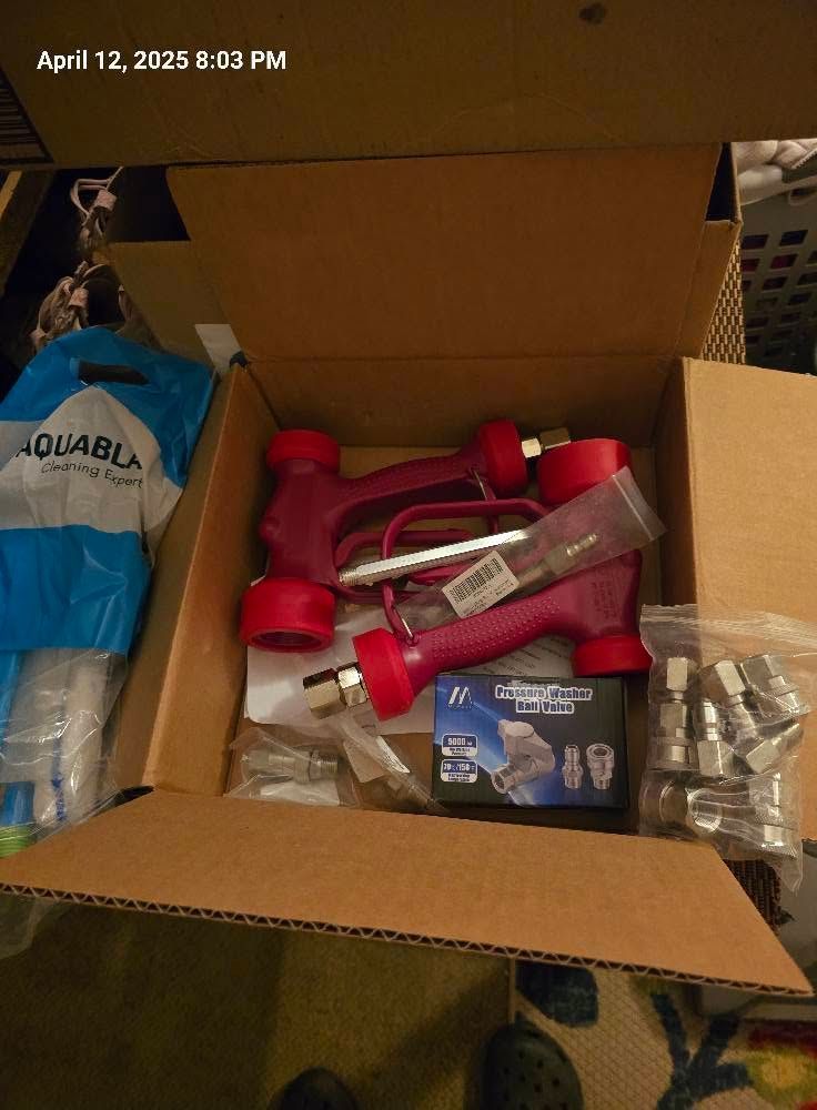 Open cardboard box with red plastic fittings, metal parts, and a blue bag on a floor.