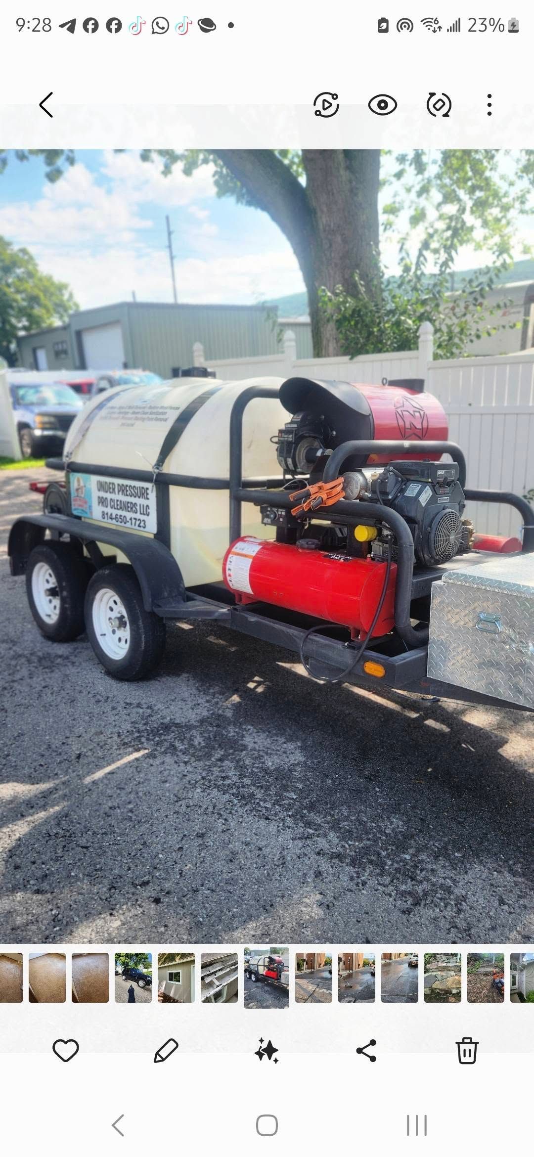 Trailer-mounted pressure washer with red engine and water tank parked on a street beside a tree