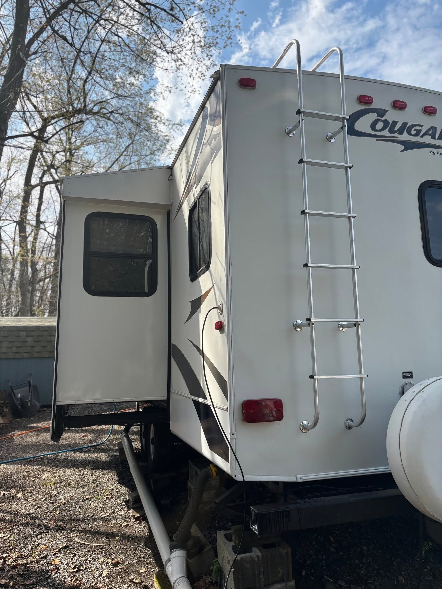 White Cougar RV with open side slide-out and rear ladder, parked outdoors among trees.