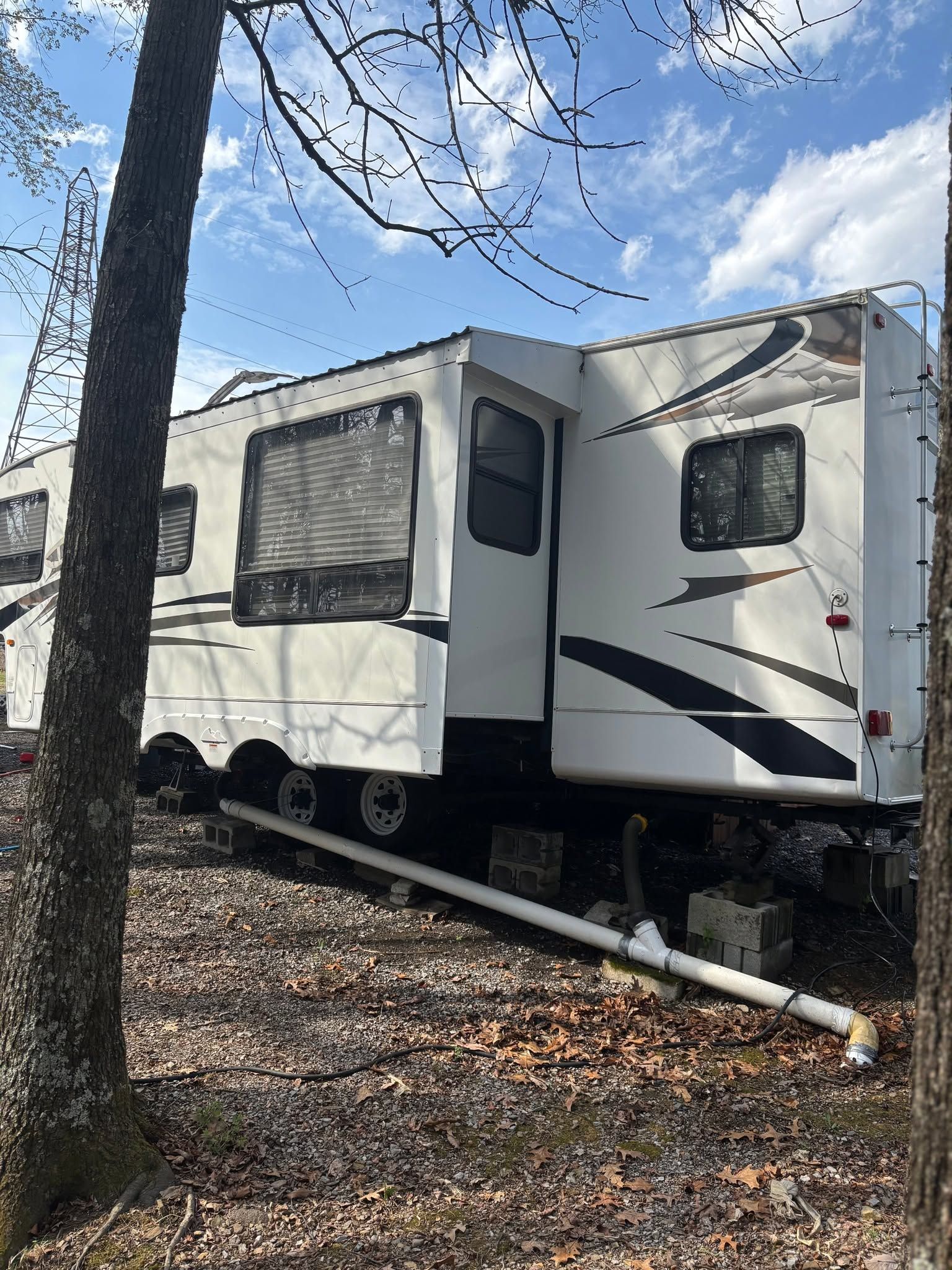 White RV trailer parked among trees on a leaf-covered slope under a partly cloudy sky