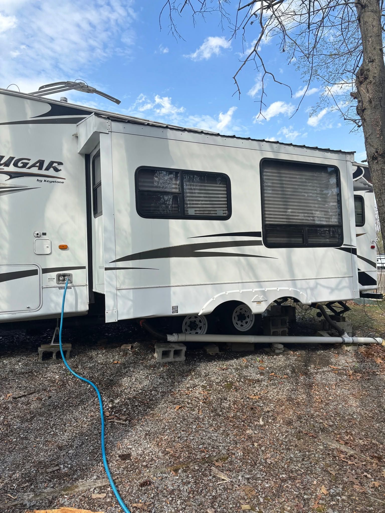 White travel trailer parked on gravel under trees, with a blue hose attached and windows closed