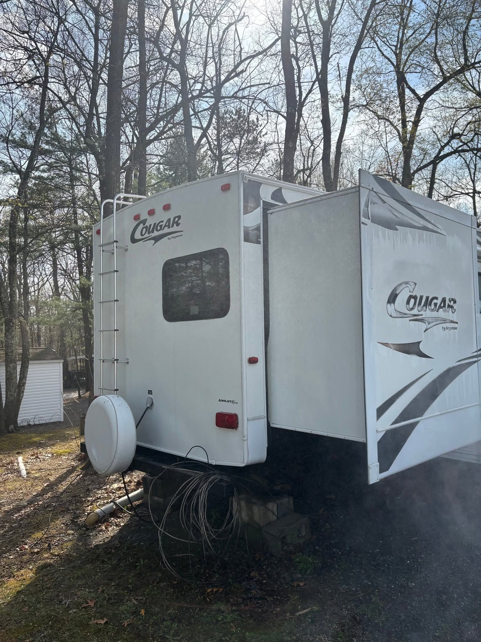 White travel trailer with slide-out parked among trees in a wooded campsite.