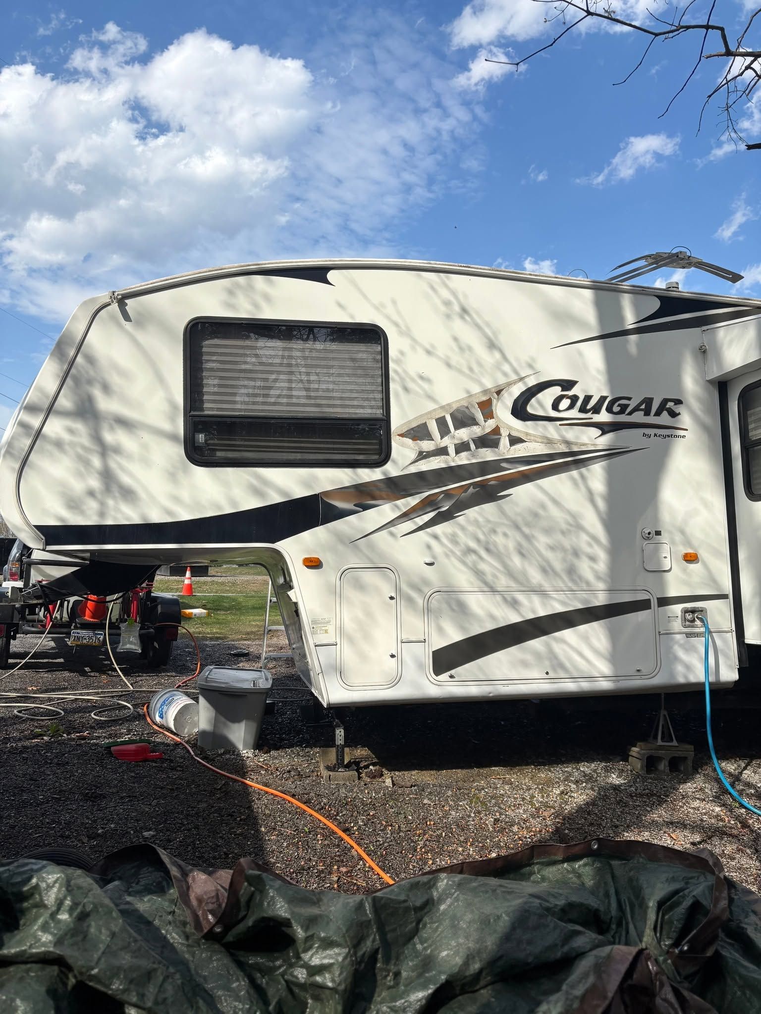 White Cougar travel trailer parked outdoors under a blue sky, with gravel and black tarps in the foreground.