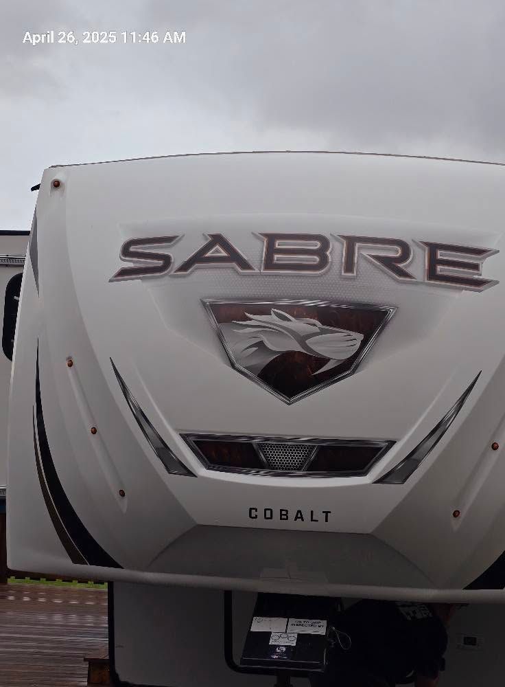 Front view of a white Sabre Cobalt RV with a bird logo and cloudy sky above
