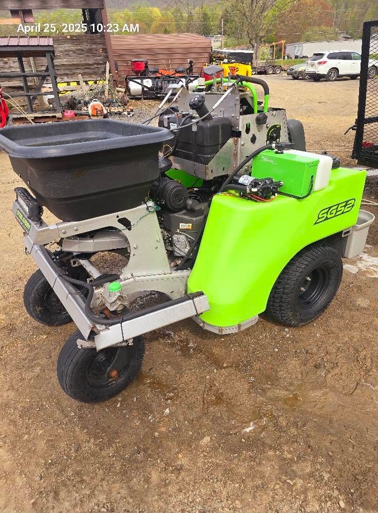 Green ride-on sweeper machine with black hopper, parked outdoors on a gravel lot