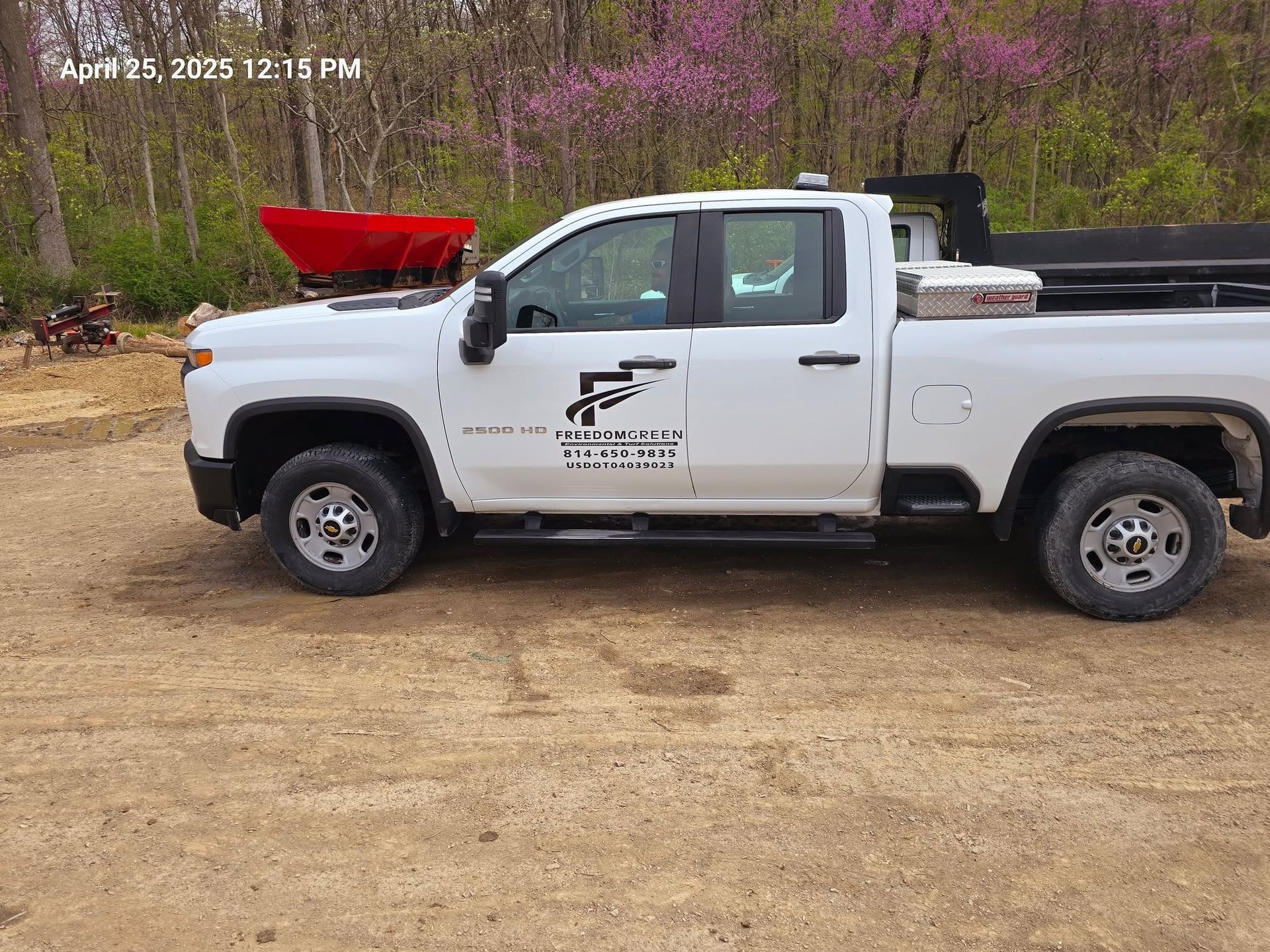 White pickup truck with a red bed-mounted hopper on a dirt lot beside trees