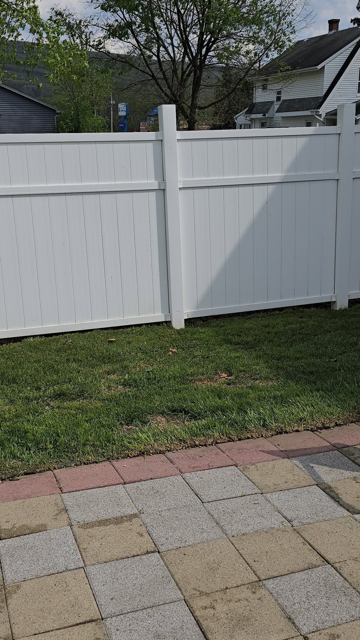 White vinyl fence with gate in a backyard, bordered by grass and a patterned patio.