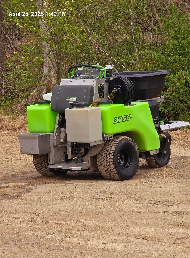 Green utility cart with a black cargo bed parked on a dirt road near trees.