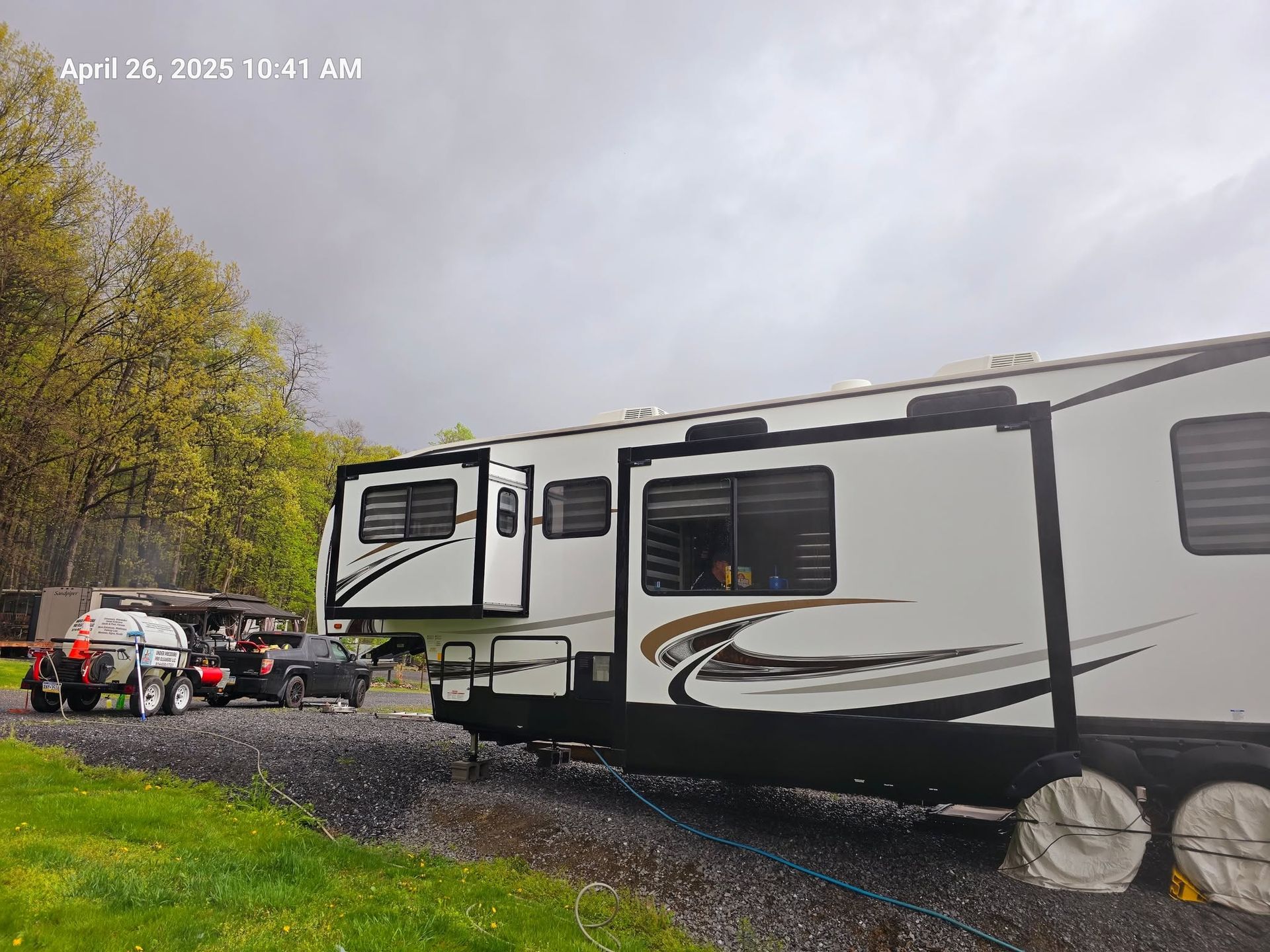 White travel trailer parked on a gravel campsite beside trees, with an awning and outdoor chairs nearby.