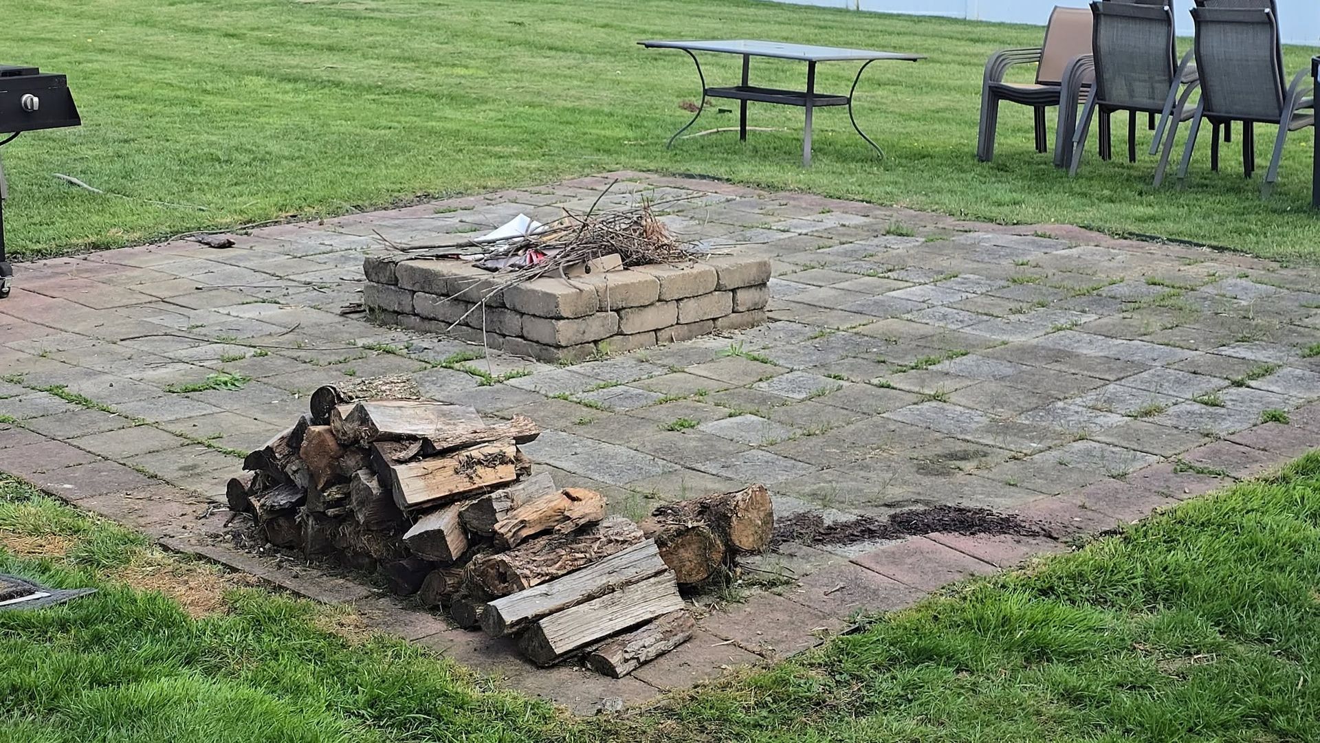 Backyard fire pit with stacked logs, surrounded by stone pavers and lawn, with picnic table and chairs nearby