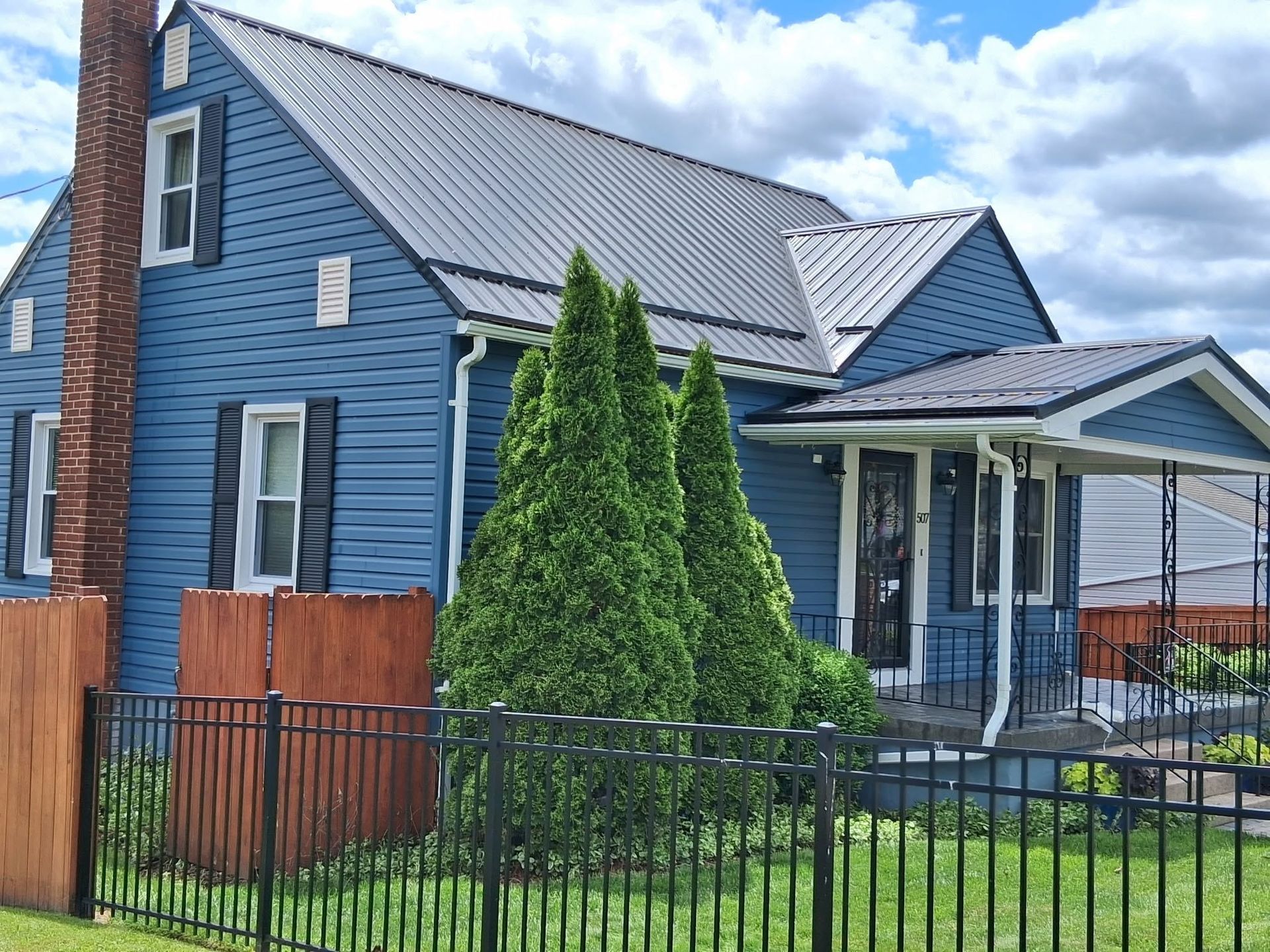 Blue house with white trim, front porch, and evergreen shrubs behind a black fence