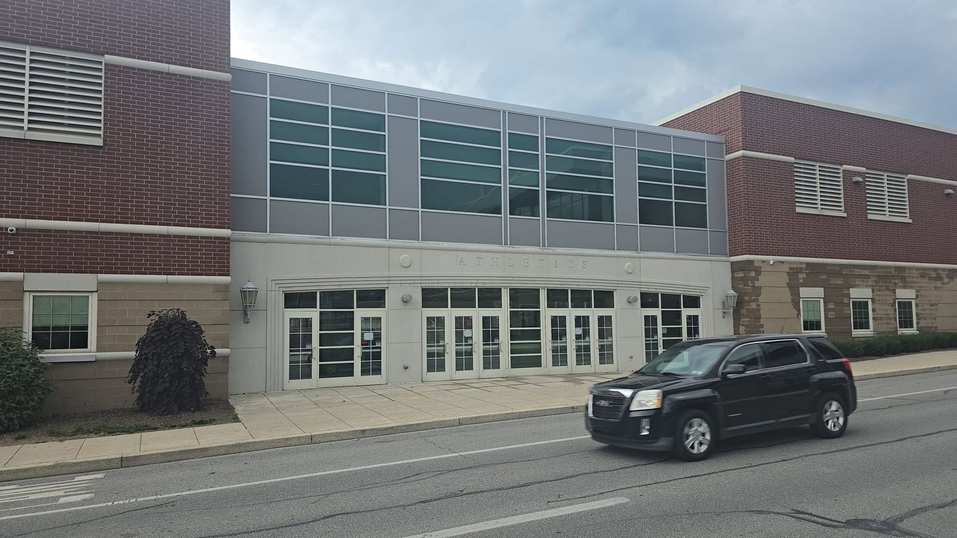 Modern building entrance with glass doors and windows, red brick facade, and a black car on the street.