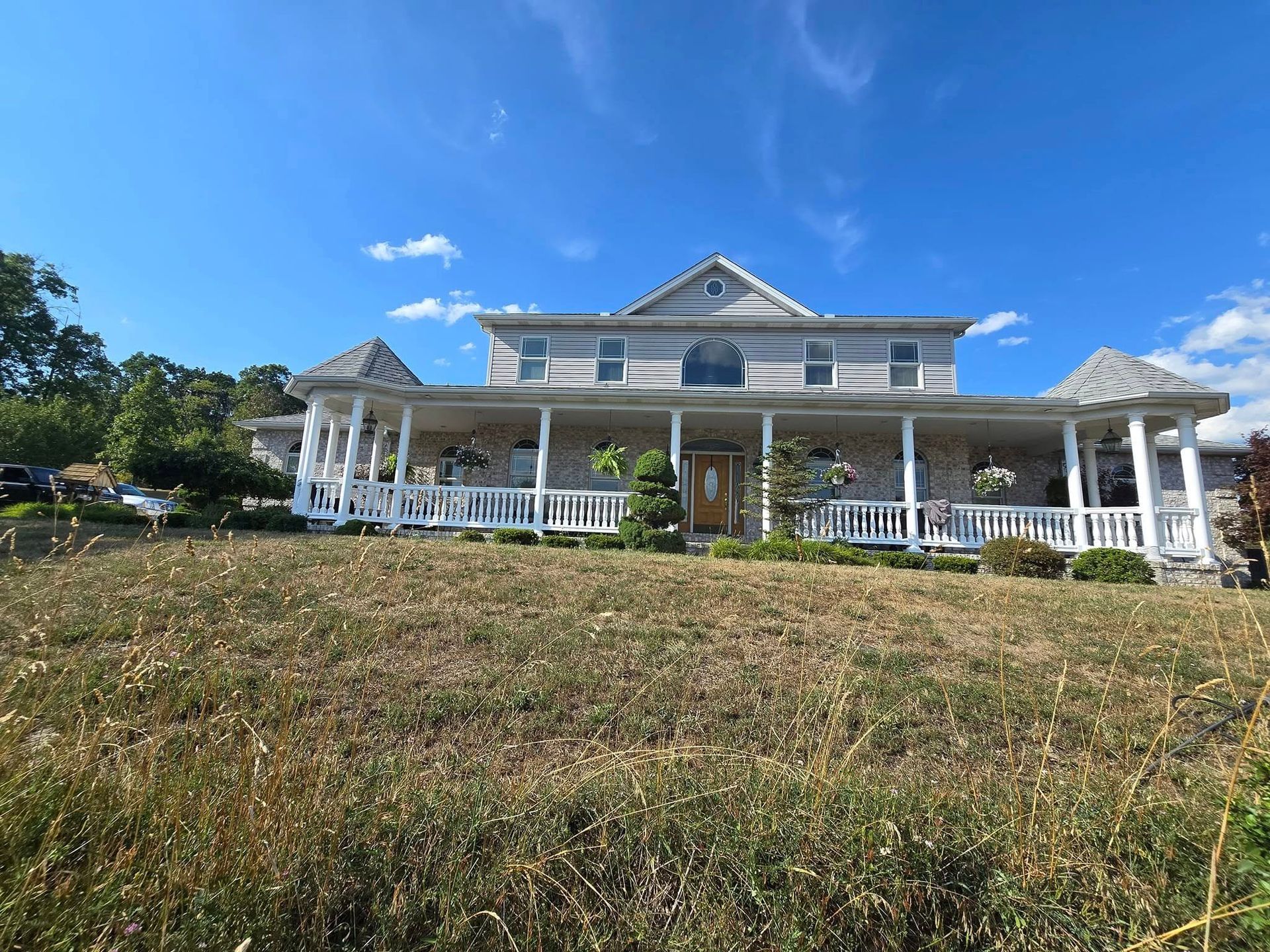 Large white two-story house with a wraparound porch on a grassy hill under a blue sky