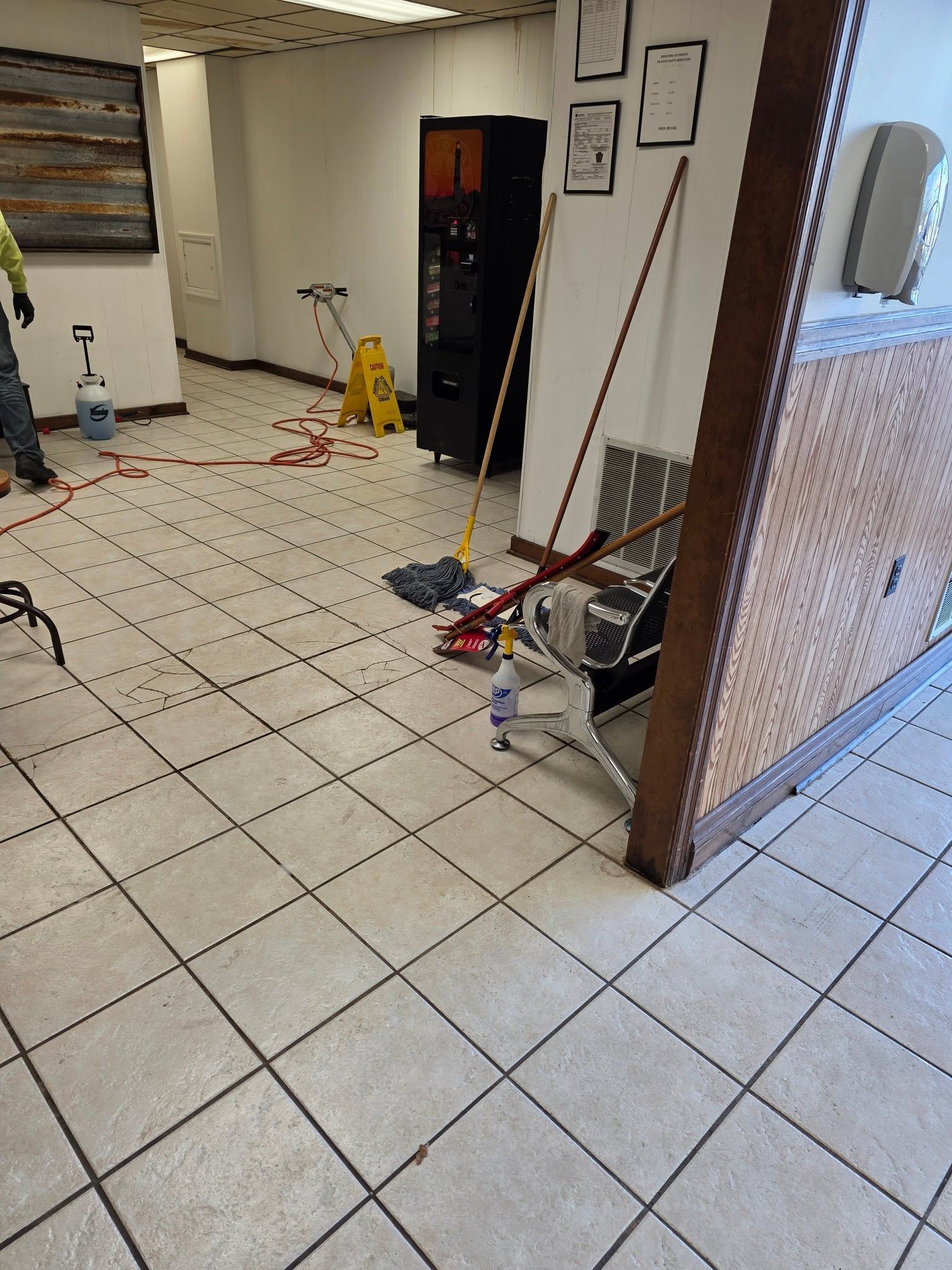 Tiled indoor lobby with a yellow caution sign, cleaning tools, and open doorway near a counter