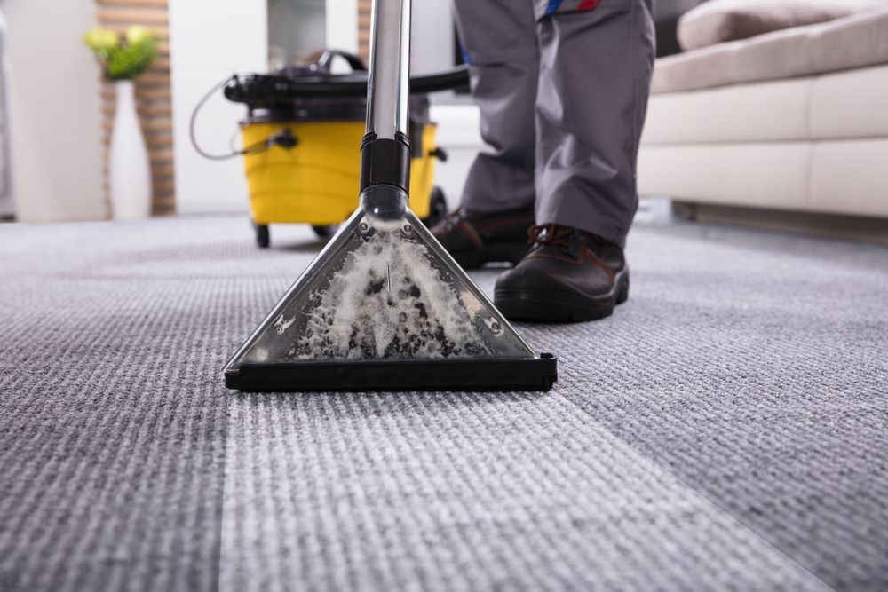 Carpet cleaner vacuuming a gray office carpet beside a yellow cleaning machine