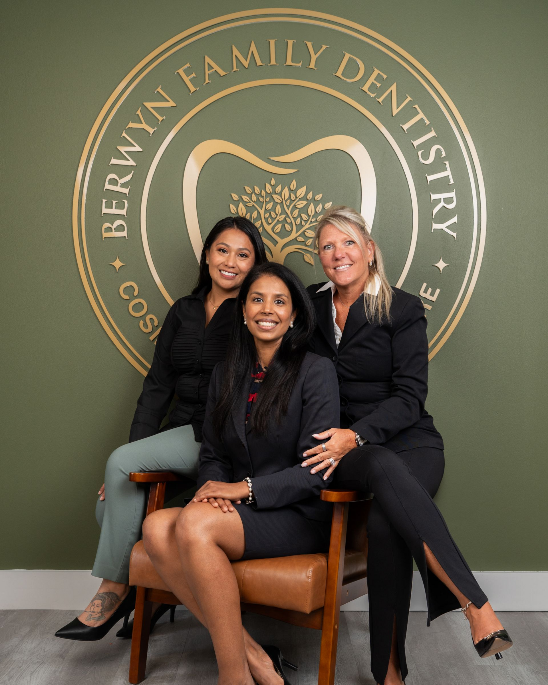 Three people in business attire pose in front of a dentistry logo on a green wall.