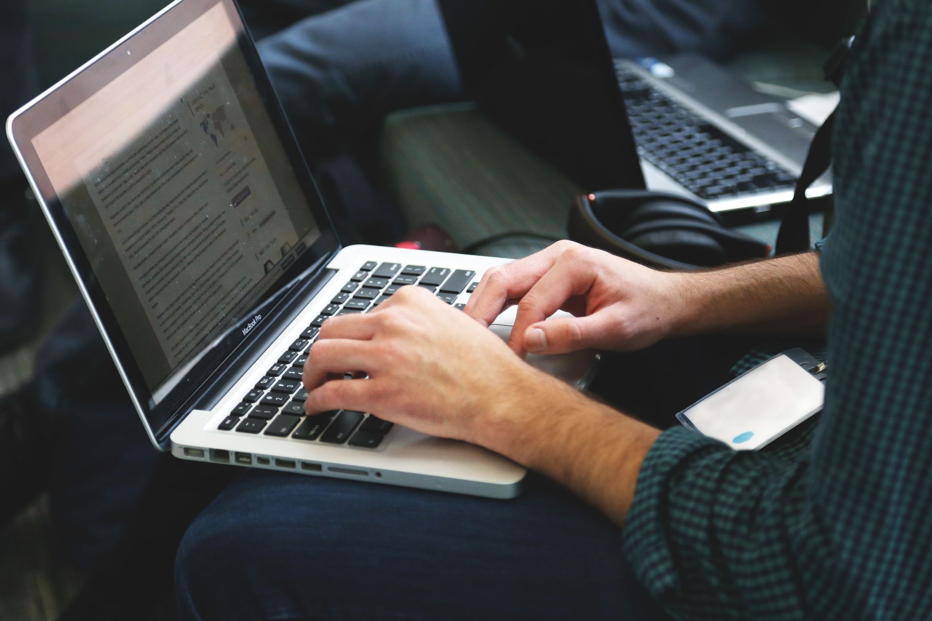Hands typing on a silver laptop while seated on a dark couch, with a second laptop nearby