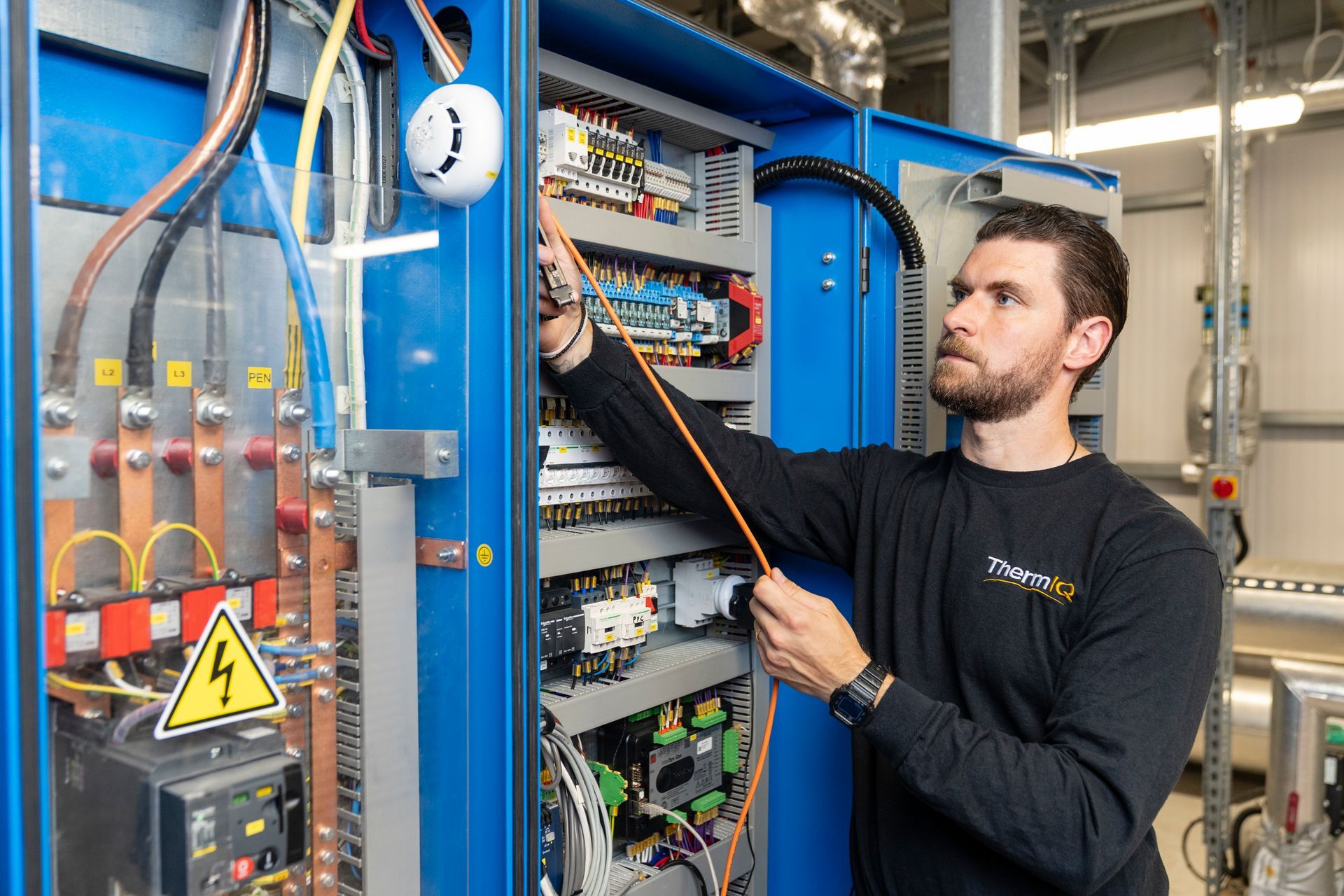 Technician inspecting electrical control panel in an industrial plant, with gauges and wiring visible.