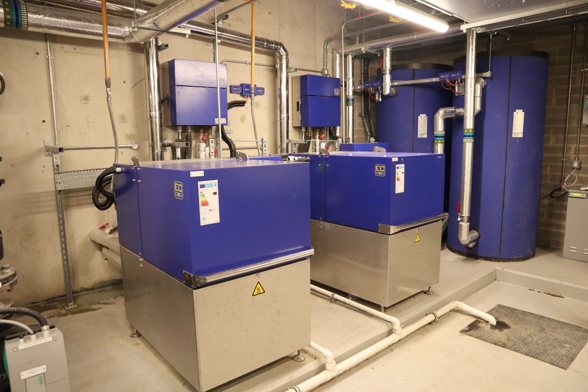 Blue industrial equipment and tanks in a mechanical room with pipes and concrete walls