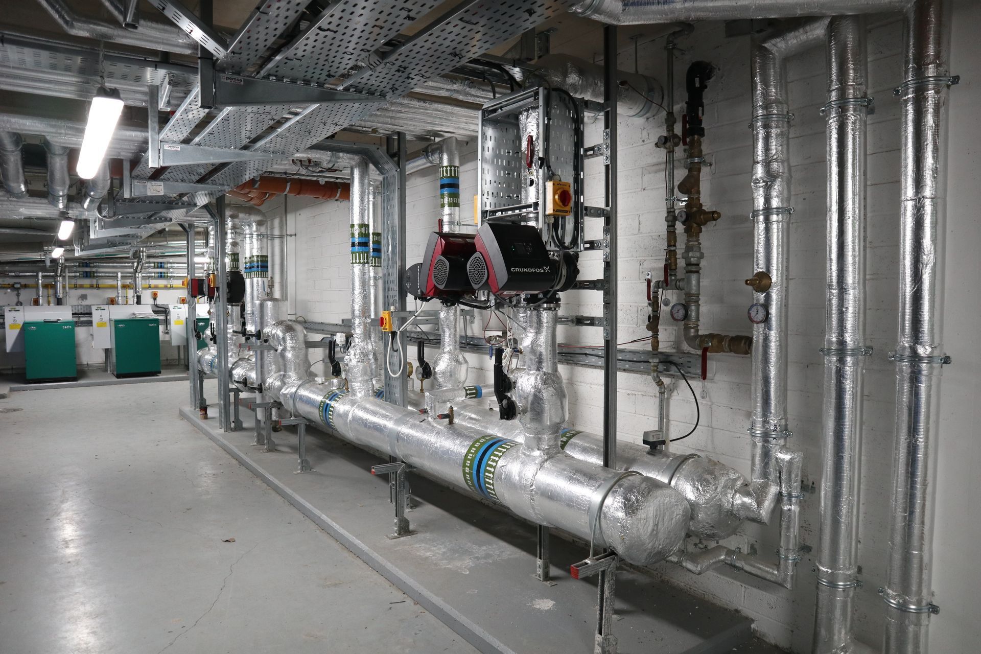 Industrial basement with large silver pipes, valves, and machinery along a concrete corridor