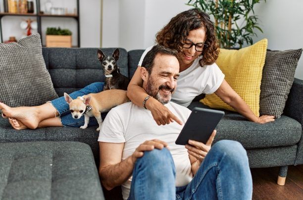 A smiling couple sitting on a couch with two small dogs, looking at a tablet together in a living room.