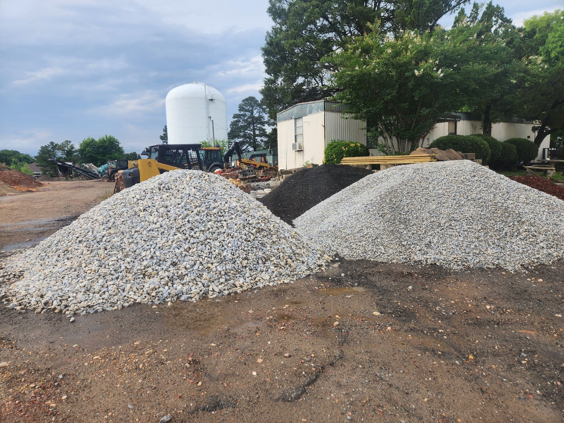 Two large gravel piles at a construction site with a water tank and trees in the background