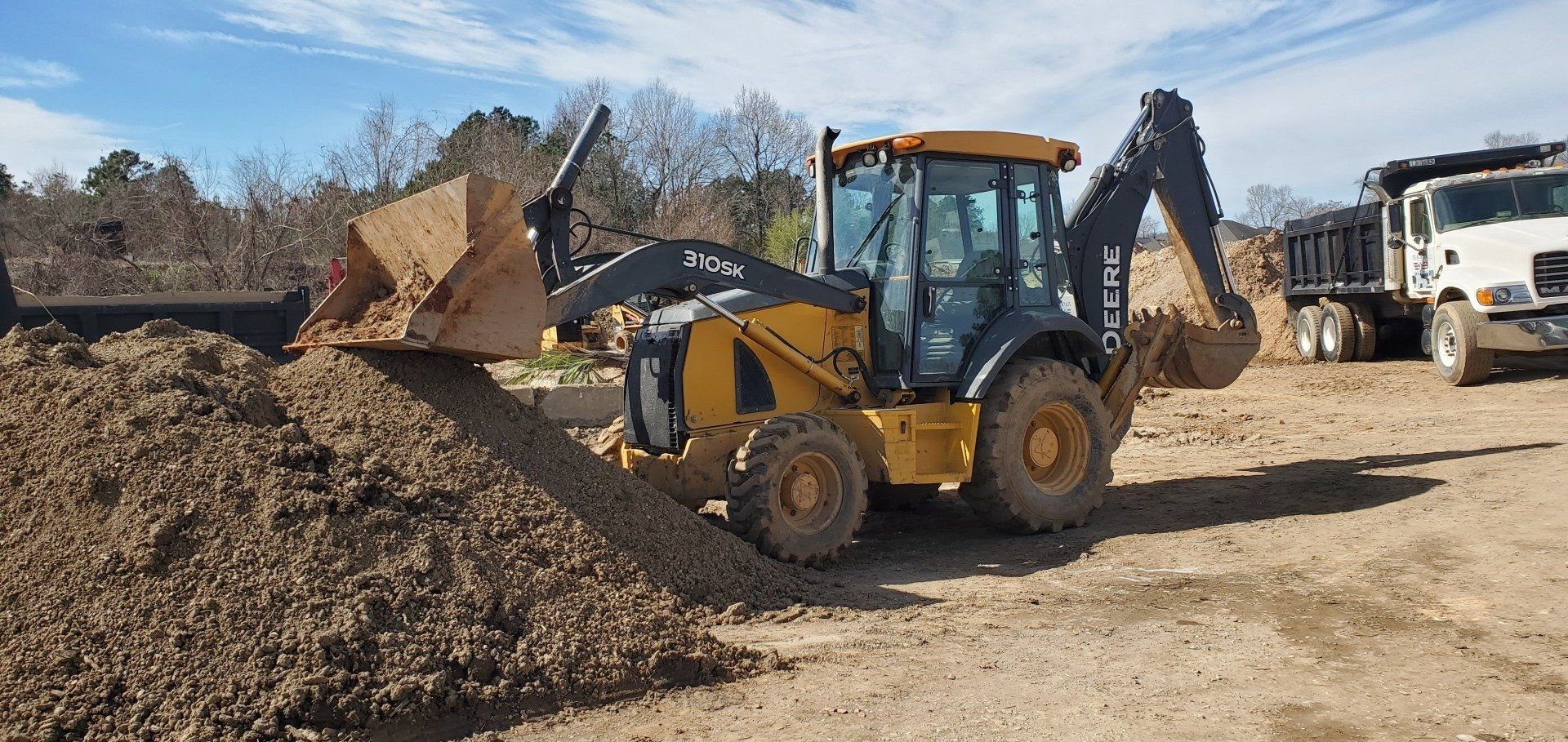 Yellow front loader moving gravel in a dirt lot, with trucks and piles of earth in the background