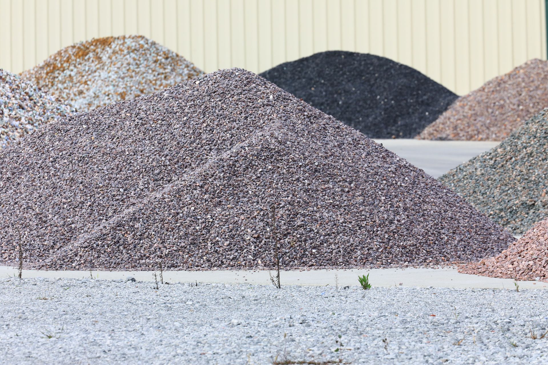 Piles of small stones and gravel in gray, brown, and pink tones stacked outdoors against a beige wall