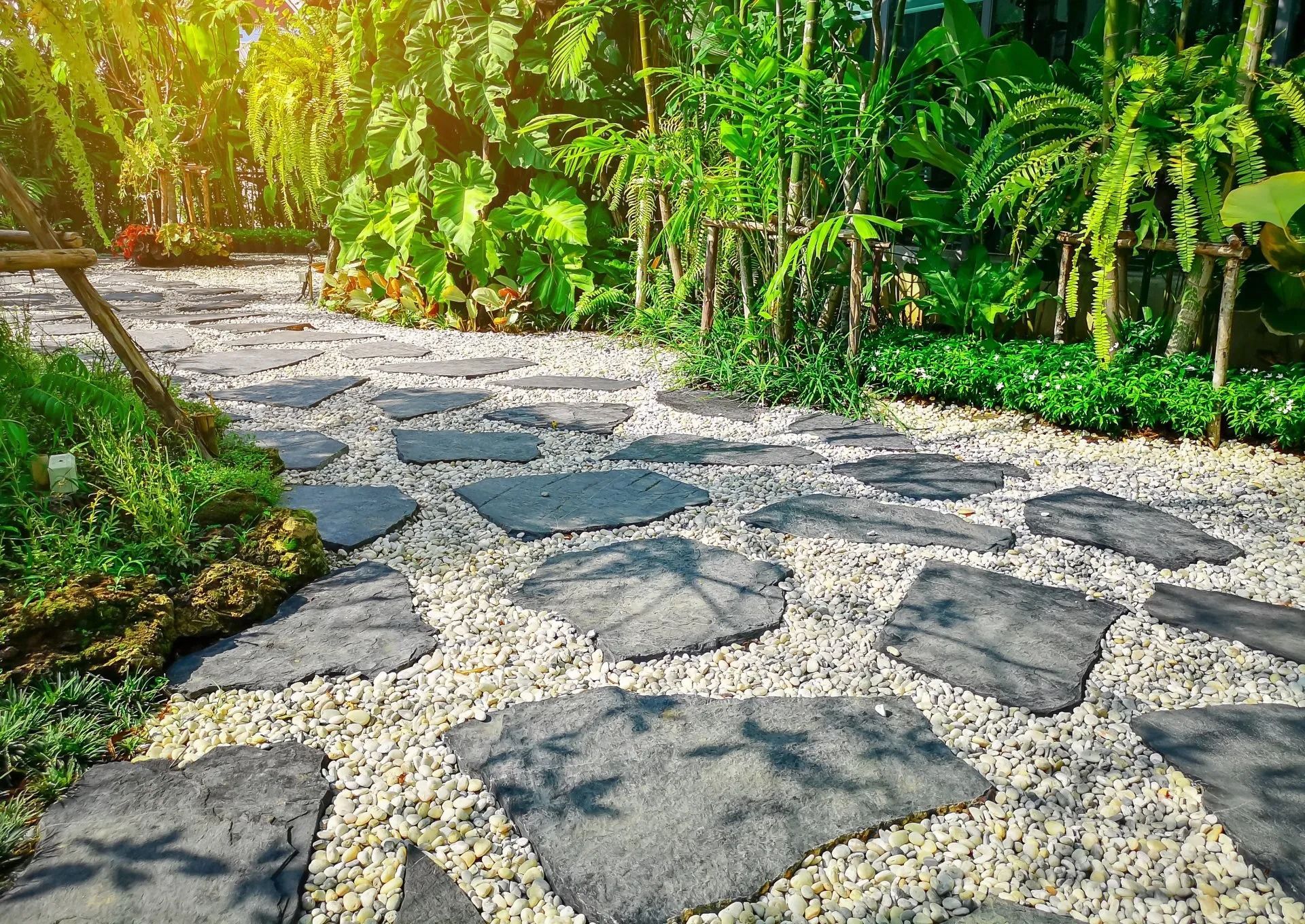 Stone pathway through lush garden, large gray stones on a bed of small white gravel, surrounded by green foliage. Stone path in garden