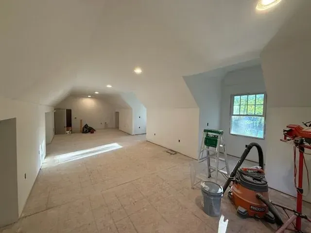 Empty unfinished room with white walls, beige floor, recessed lights, and painting supplies near a window