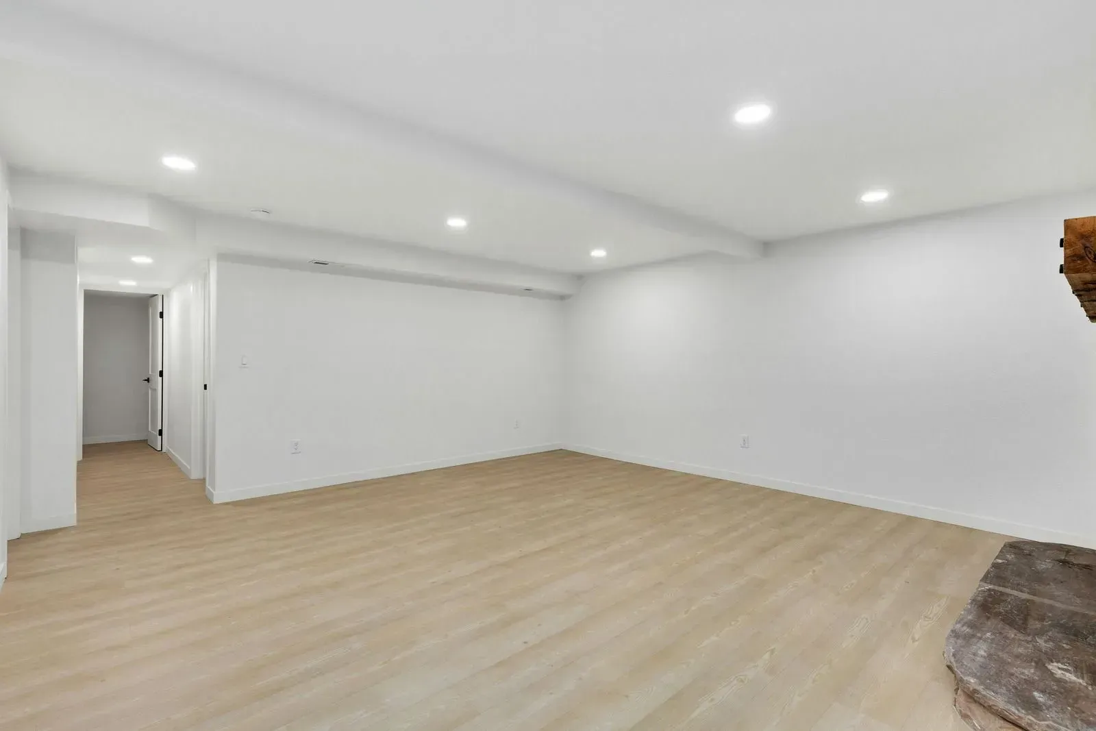 Empty white room with light wood floors, recessed ceiling lights, and a doorway hall on the left.