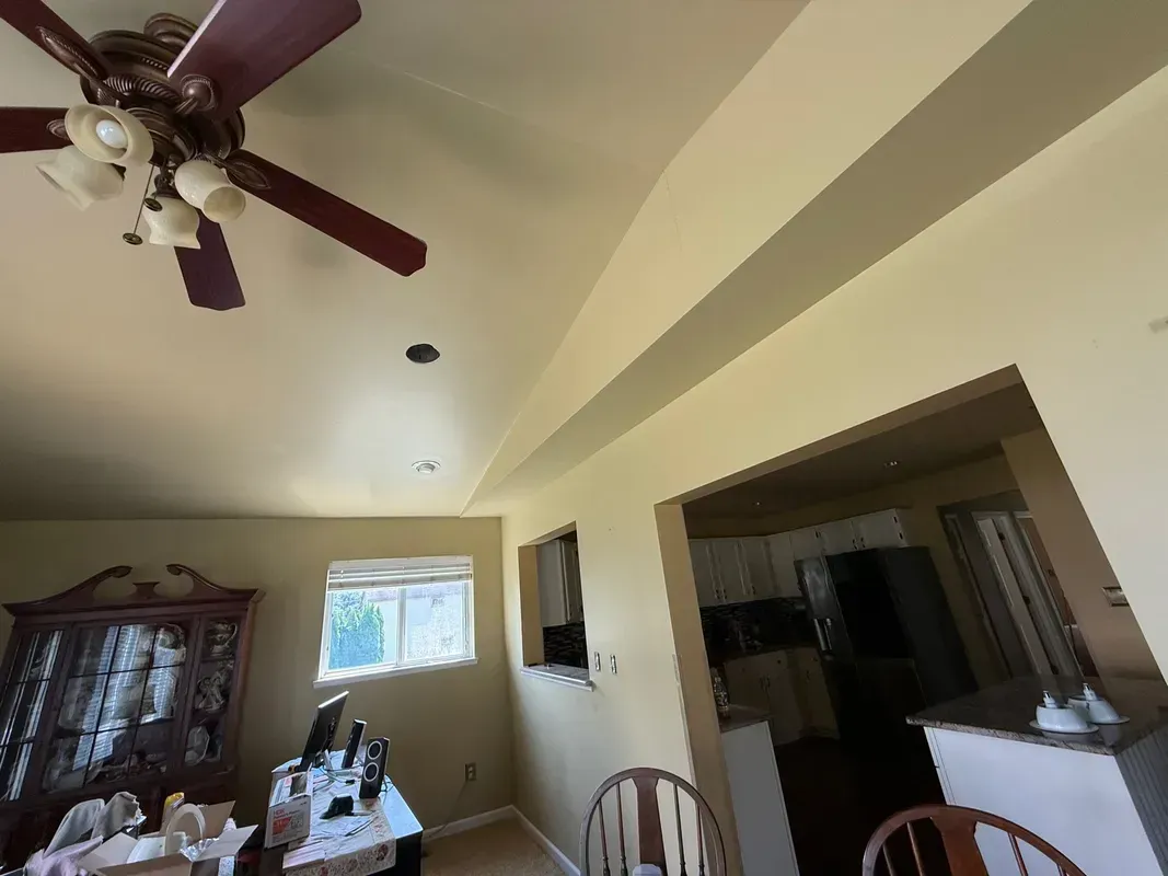 Dining room with ceiling fan, wooden table, and cabinet, viewed into a kitchen through a wide doorway.
