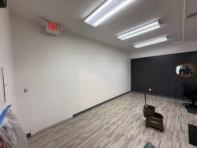 Empty office room with white walls, gray wood floor, black back wall, and fluorescent ceiling lights.