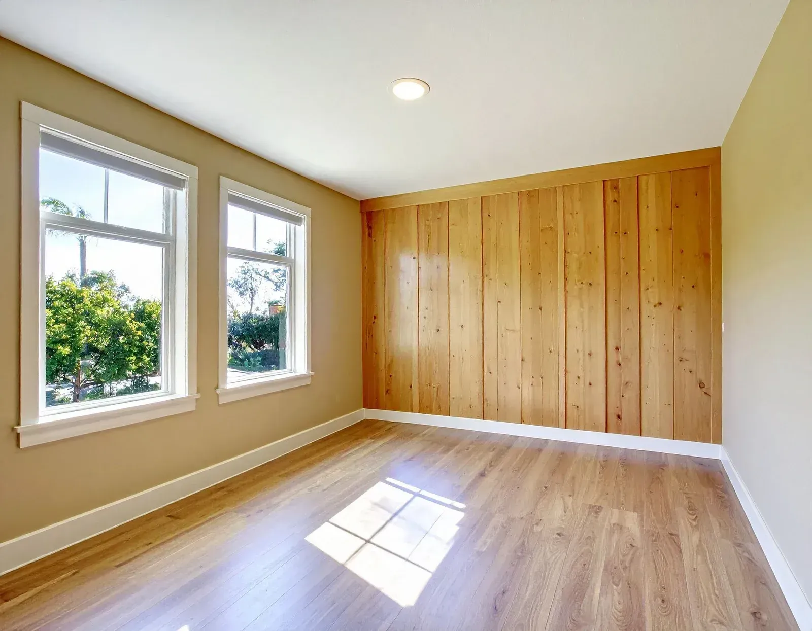 Bright empty bedroom with hardwood floor, beige walls, wood accent wall, and two windows